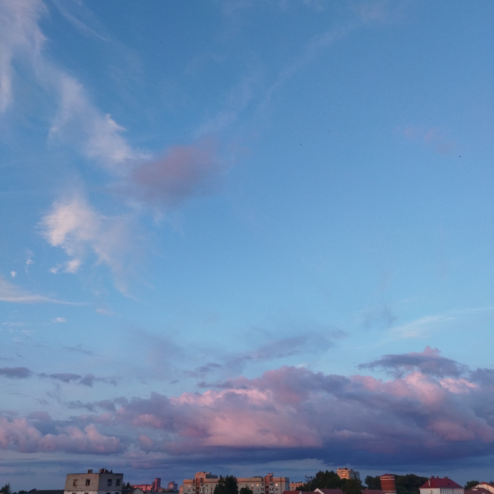 This is a photograph of a pale blue sky with wispy white and pink clouds at sunset, seen from a rooftop in Polatsk, Belarus. The lower third of the image shows the tops of various buildings, including a light-colored apartment building with visible windows and a darker red-tiled roof. The sky is a dominant feature, with softer hues of pink and purple coloring some of the clouds, suggesting the transition from day to night. There are no people or discernible actions visible in the scene. The environment appears to be urban, with the architecture of the buildings indicating a European setting. The photograph is taken from a high vantage point, looking towards the horizon.