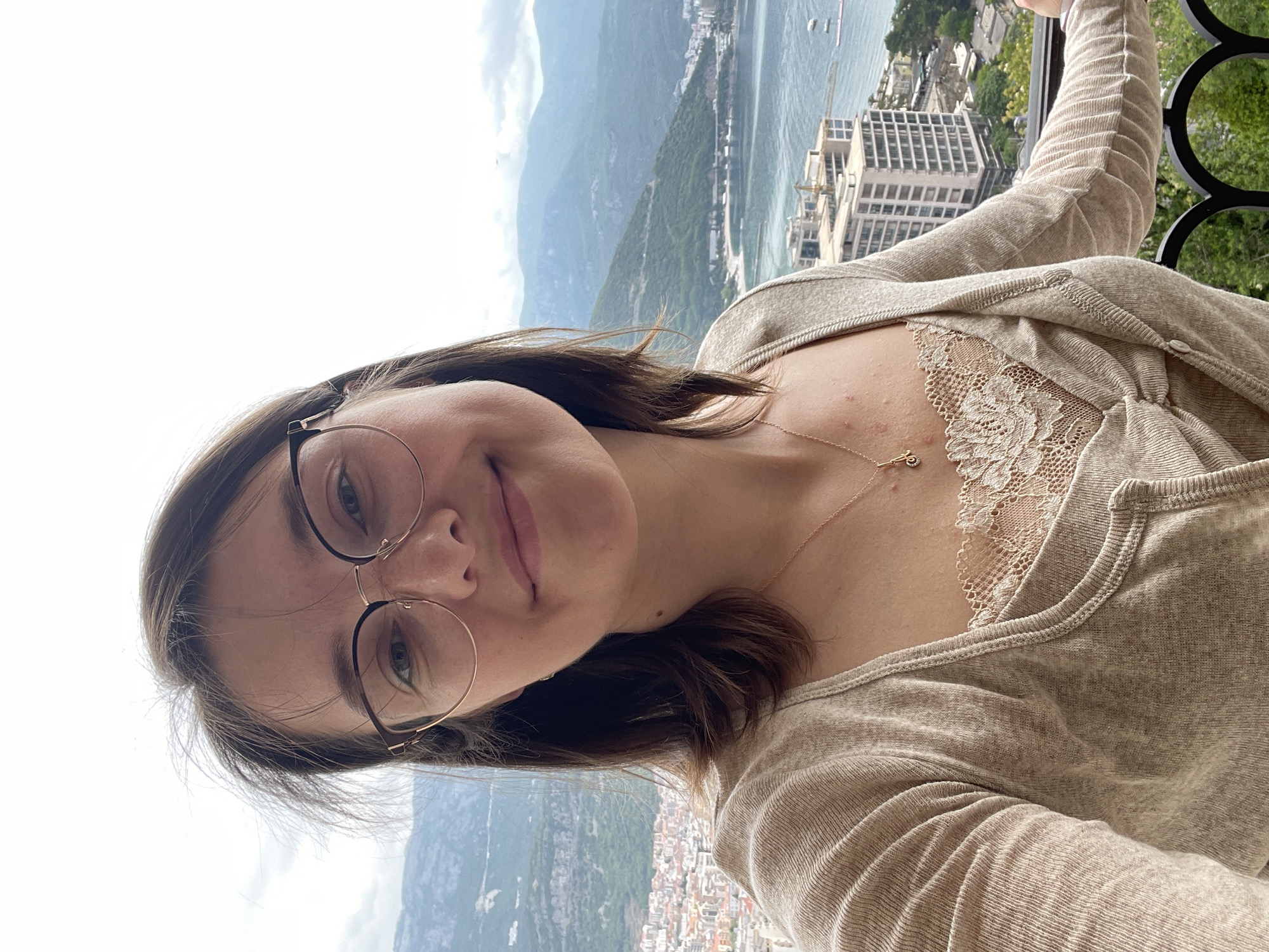 A woman wearing glasses, a beige cardigan, and a lace-trimmed top is pictured from a close-up, slightly tilted angle. She is on a balcony with a wrought-iron railing, which is visible at the top right. Behind her, the city of Budva, Montenegro is visible, showing buildings near the coast and mountains in the distance. The sea is visible between the mountains and the city.