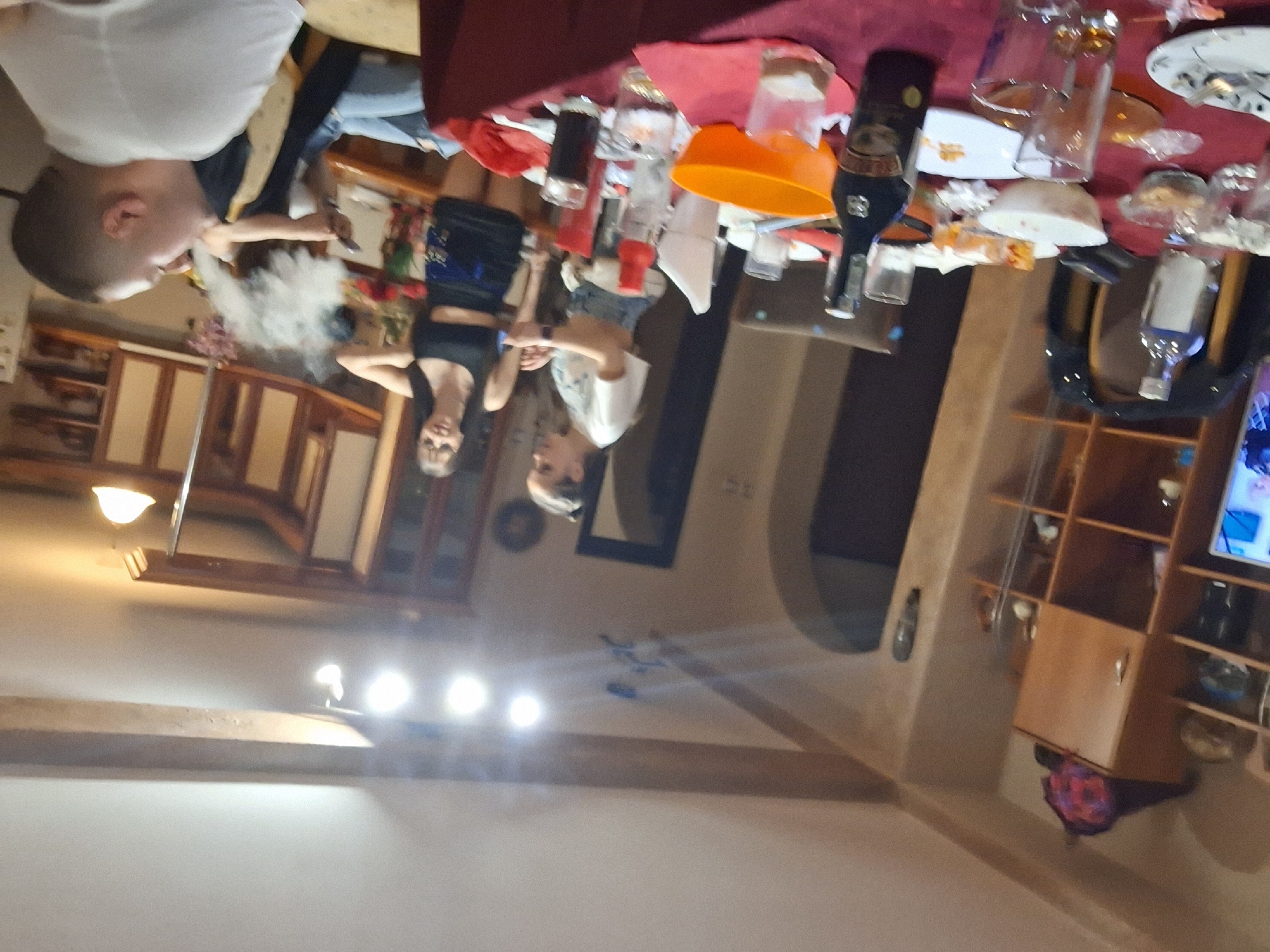 The image shows an indoor scene, likely a home dining room or living room. The focus is on a table covered with a red tablecloth and various items, including plates, glasses, bottles of alcohol, and an orange bowl. Three individuals are visible around the table, two women standing and a man sitting. The man is exhaling a large amount of vapor. A television and a display shelf with items can be seen in the background. The location cannot be determined as being in the city of Panagyurishte, Bulgaria.