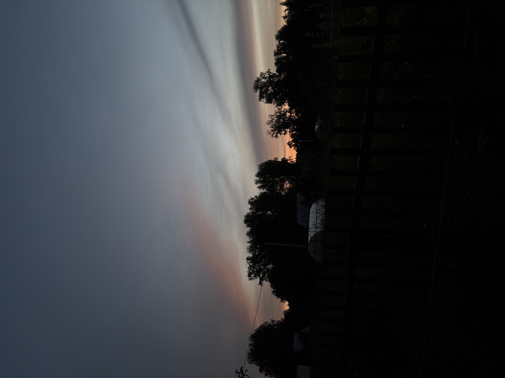 The image shows a sunset over a rural area, likely a village or dacha settlement, in Dubrovino, Russia. The sky is a gradient of dark blue at the top, transitioning to lighter blues and grays, with streaks of orange and pink from the setting sun. Silhouetted against the sky are a line of trees and some structures, possibly greenhouses, with a dark wooden fence in the foreground. The scene is dim, suggesting it is late evening or early morning. There are no people or specific actions depicted.