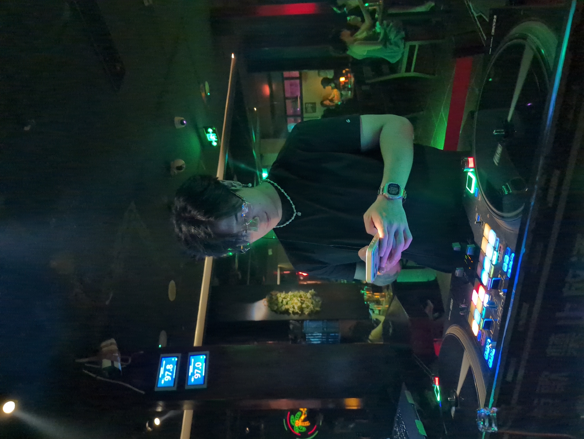 In a dimly lit venue in Liangjiang, China, a DJ stands behind a mixing console illuminated with blue and red lights. The DJ, wearing glasses, a black shirt, and a watch, holds a smartphone in their hand. A green hue dominates the background, where other people can be seen seated at tables. Indicators reading "97.0" are visible, possibly referencing frequencies. A display of snacks sits on the counter in front of the DJ.
