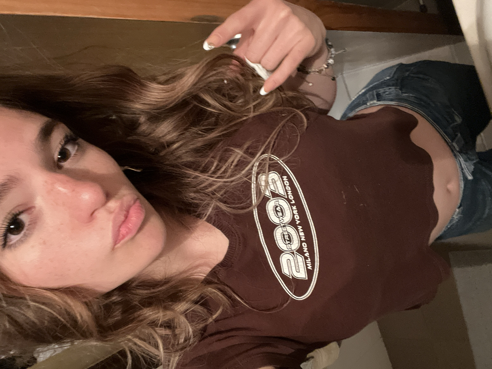 A young woman with long, wavy brown hair and light skin is taking a selfie. She is wearing a brown crop top with white lettering and blue jeans. Her fingernails are painted white. She is holding up her hand, making a gesture with her fingers. The background is out of focus. It is impossible to identify the location as San Bernardino Tlaxcalancingo, Mexico, from the image alone.