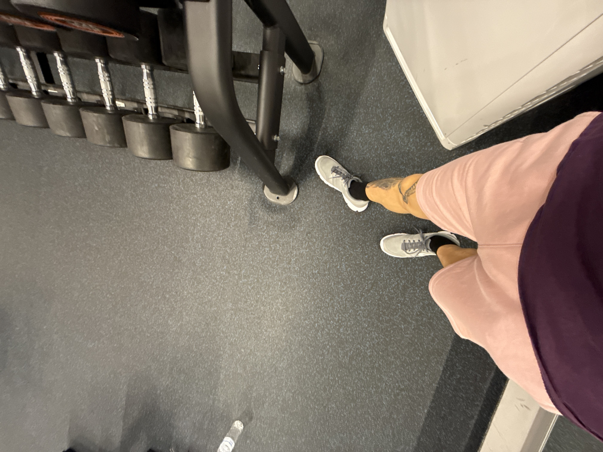 The image appears to be taken from an elevated first-person perspective, looking down at a gym setting. To the left, there is a dumbbell rack loaded with a set of black dumbbells. The flooring is a dark, textured rubber material. Towards the right side of the image is a white, rectangular object that could be part of a fitness machine or storage unit. A person is standing on the right, visible from the ankles down. The person is wearing grey athletic shoes with black socks and has tattoos on the left ankle. They are also wearing light pink shorts and a dark purple top or shirt. A water bottle is visible near the bottom-left corner of the image. Based on the presence of gym equipment, flooring, and clothing, the location is likely inside a fitness center. The image does not provide any information that indicates the city of Sofia, Bulgaria.