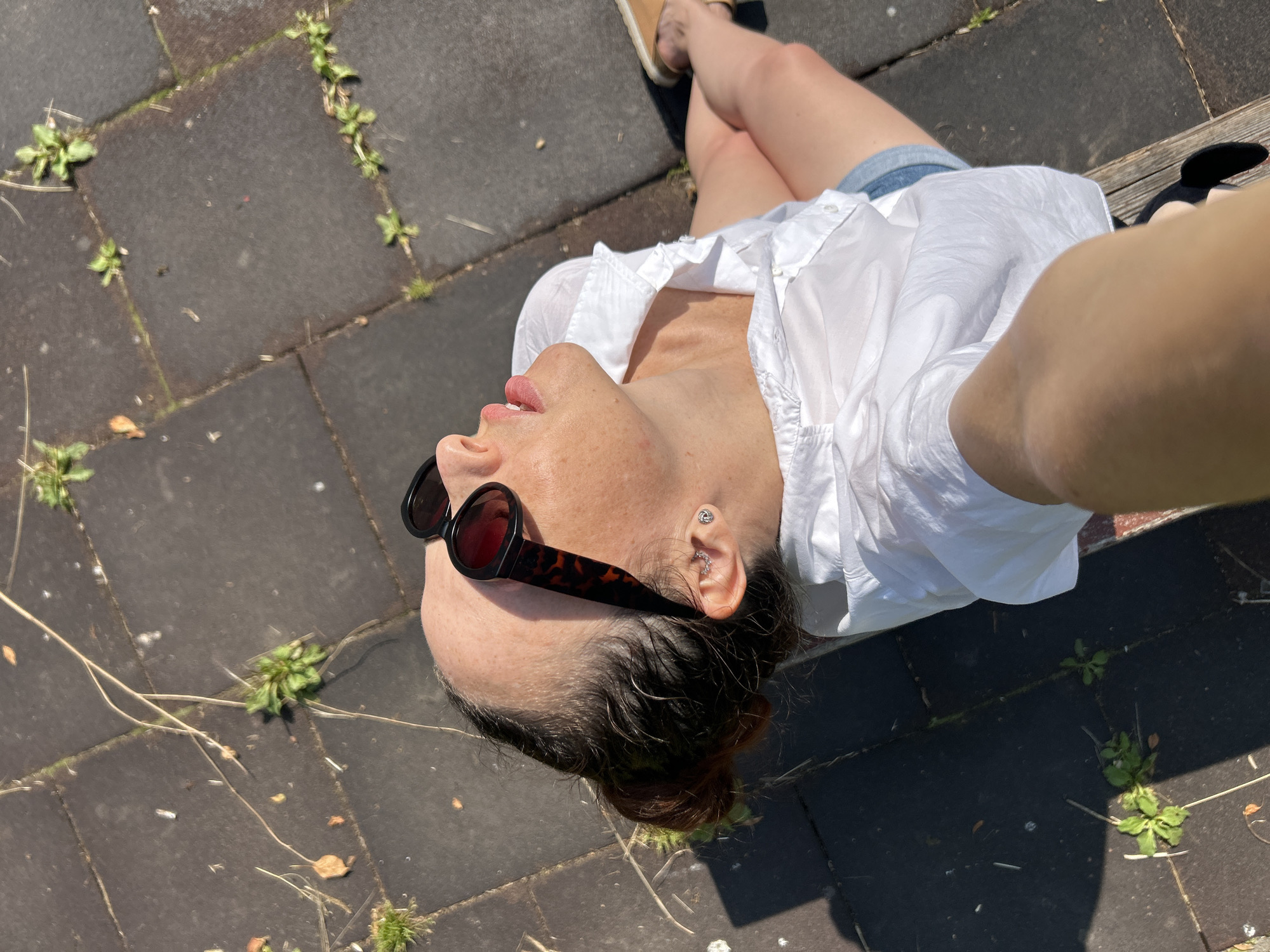 A woman is sitting on a wooden bench outdoors on a sunny day, looking up at the sky. She is wearing a white collared shirt and denim shorts. She has on dark-rimmed sunglasses and a silver earring in her left ear. Her hair is pulled back in a bun. The ground is paved with dark square tiles, with small weeds growing in the cracks between them. The scene appears to be in a park or public area.