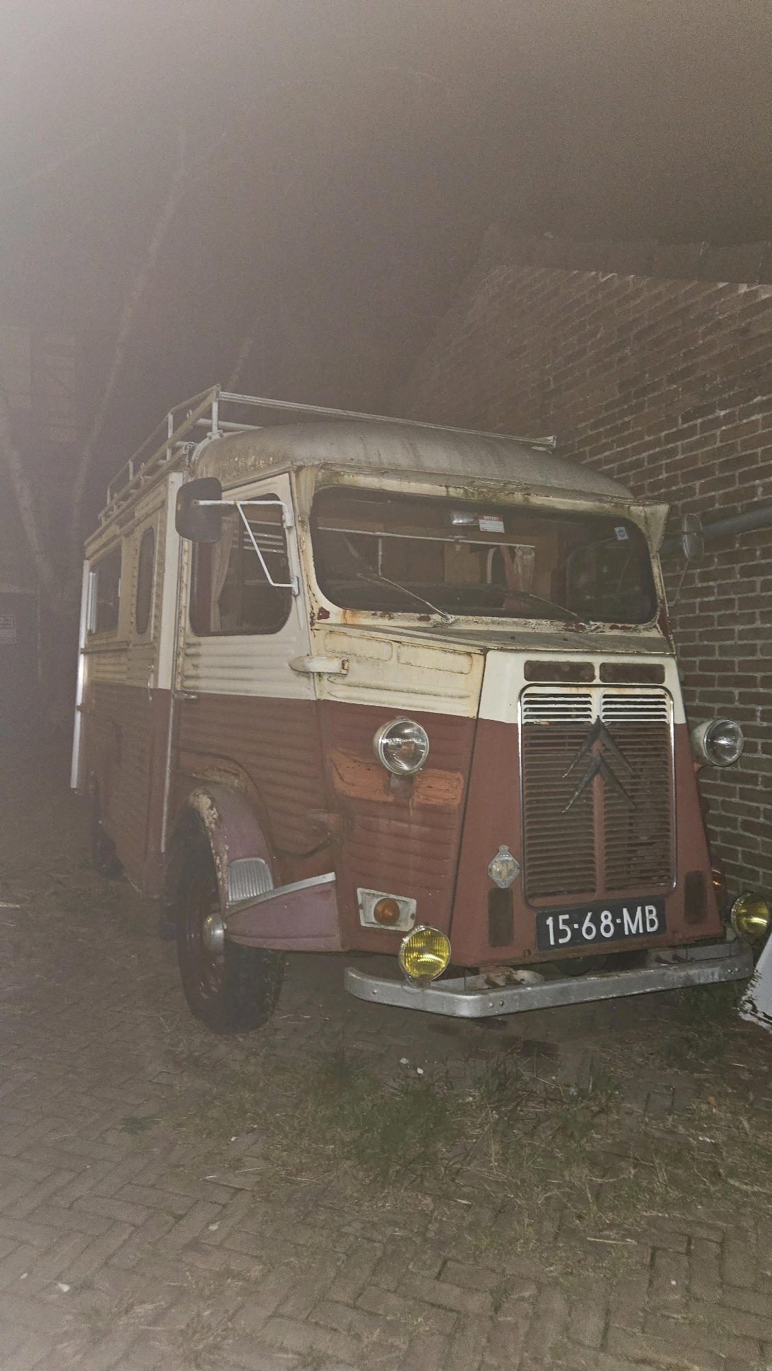 The image shows a vintage, two-tone red and white van parked on a brick-paved surface, possibly a driveway or alleyway, adjacent to a red brick wall. The van, a Citroën H Van, is weathered, with visible rust and a roof rack. It has a license plate that reads "15-68-MB". The scene appears to be outdoors and dimly lit, with a hazy or foggy atmosphere present, particularly in the background. Trees are partially visible to the left of the van. The overall condition of the van suggests it may be unused or undergoing restoration. There are no people visible in the image. The location is identified as being in the city of Breda, Netherlands.