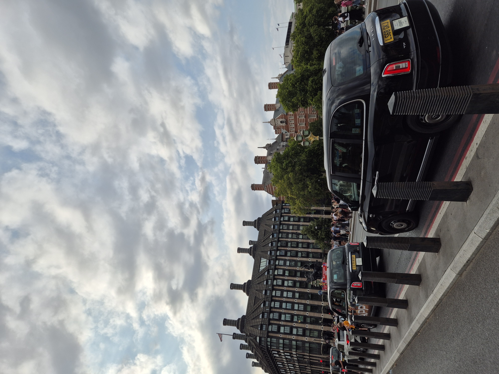 The image captures a view of the South Bank of the River Thames in London, United Kingdom, with the Houses of Parliament and Big Ben visible in the background. The foreground features a street scene with several black London taxis, commonly known as "black cabs," in motion. One taxi in the center has a license plate that reads "LS68 GKJ". The street is lined with bollards, and pedestrians can be seen walking along the pavement. The architecture in the background is characteristic of Victorian-era buildings, with distinctive red brickwork and ornate details. The sky is overcast with dramatic clouds, suggesting a typical London weather day. The scene is busy with traffic and people, indicating a vibrant urban environment.