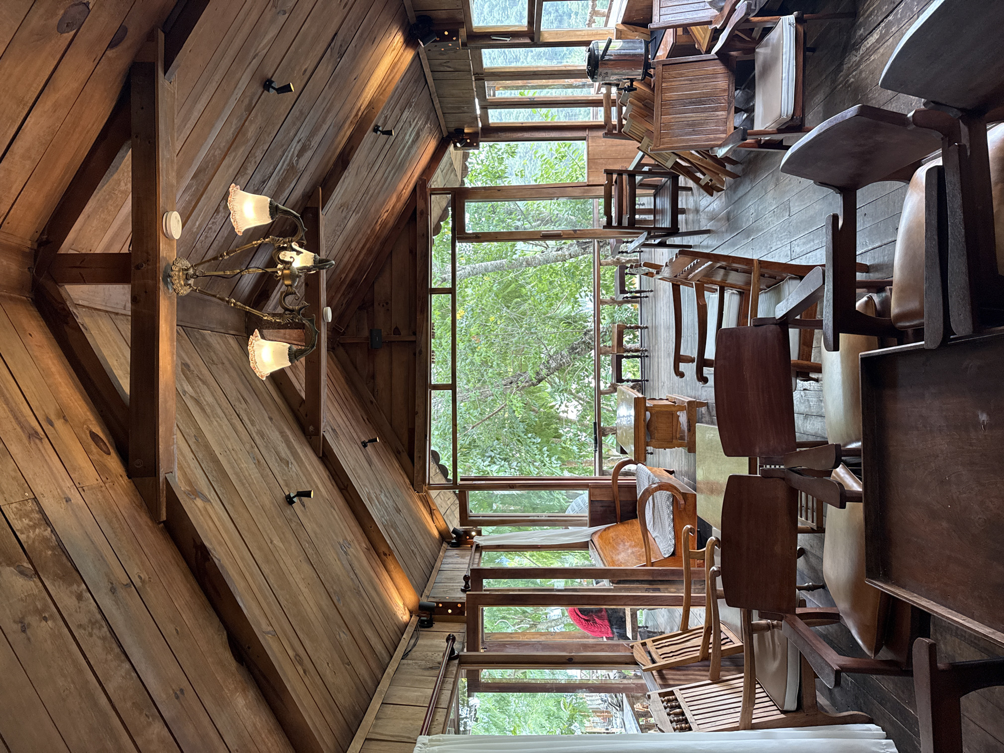 This is an interior shot of a wooden cafe with an open-air feel, located in Da Lat, Vietnam. The cafe features a rustic, natural aesthetic with extensive use of wood for the ceiling, walls, floors, and furniture. The ceiling is a sloped, planked wood structure with exposed beams, supporting a chandelier with multiple lamps. Several modern track lights are also visible on the ceiling.
The cafe is furnished with various wooden chairs and benches, some with cushions. In the foreground, a low wooden table is prominent, with another similar table and chairs further back. A wooden sofa with a patterned cushion is situated against the back wall. The cafe is surrounded by large windows offering a view of lush green trees and foliage, suggesting it is located in a natural setting, possibly a hillside or a forested area within Da Lat. The overall atmosphere is cozy and inviting, with warm lighting.