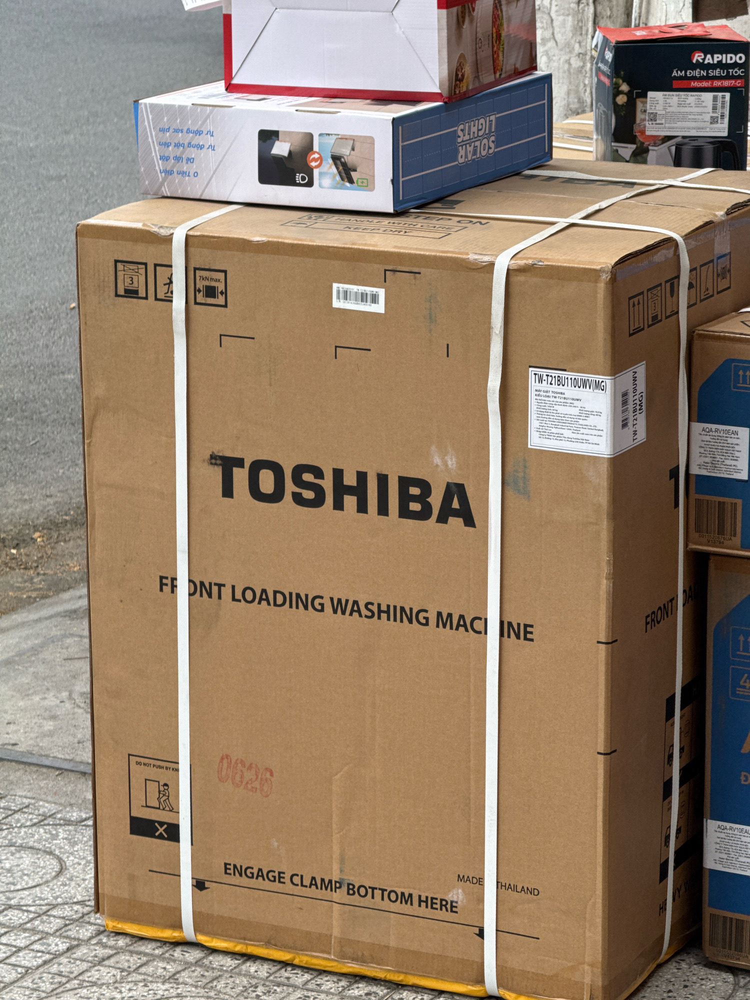 The image depicts a collection of cardboard boxes, likely containing appliances, stacked on a paved surface. The primary focus is a large, brown cardboard box prominently displaying the "TOSHIBA" logo in black lettering. Below the logo, the text "FRONT LOADING WASHING MACHINE" is visible. This box is secured with white strapping tape. Other boxes are visible around it, some featuring smaller text and labels. One box on the right has blue accents and text, including "AQA-RV10EAN". Another box on the top left has text in Vietnamese. The scene is set outdoors, with a grey, speckled paved surface in the foreground and hints of a street or sidewalk. The overall impression is of a delivery or storage area for new goods. The lighting suggests it is daytime.