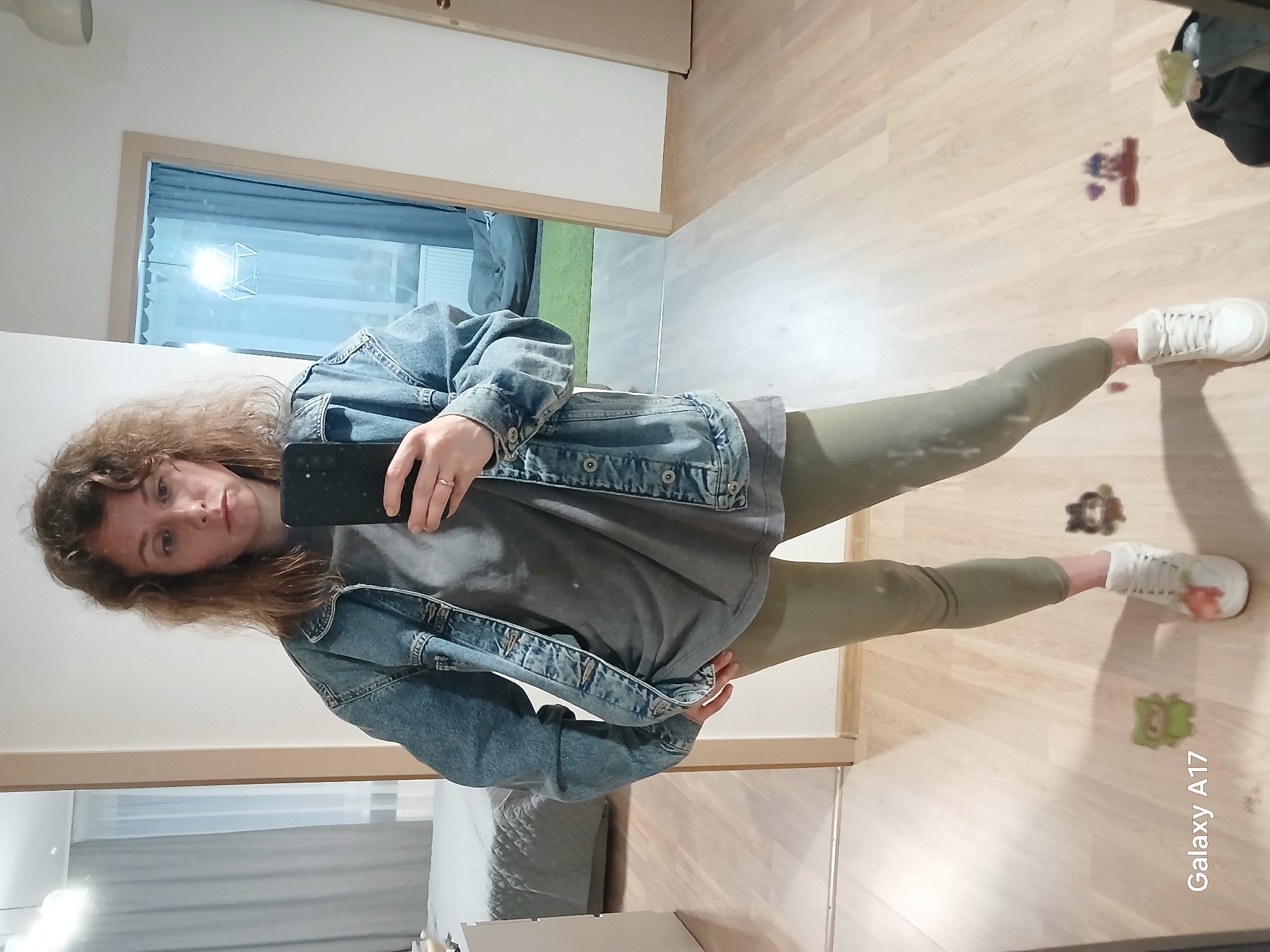A woman is taking a mirror selfie in a room with light wood flooring. She has curly brown hair and is wearing a denim jacket over a gray shirt and olive-green leggings with white sneakers. The photo appears to have been taken using a Samsung Galaxy A17. The background includes a doorway leading to another room with a grey sofa. The room is within a residence in Yekaterinburg, Russia.
