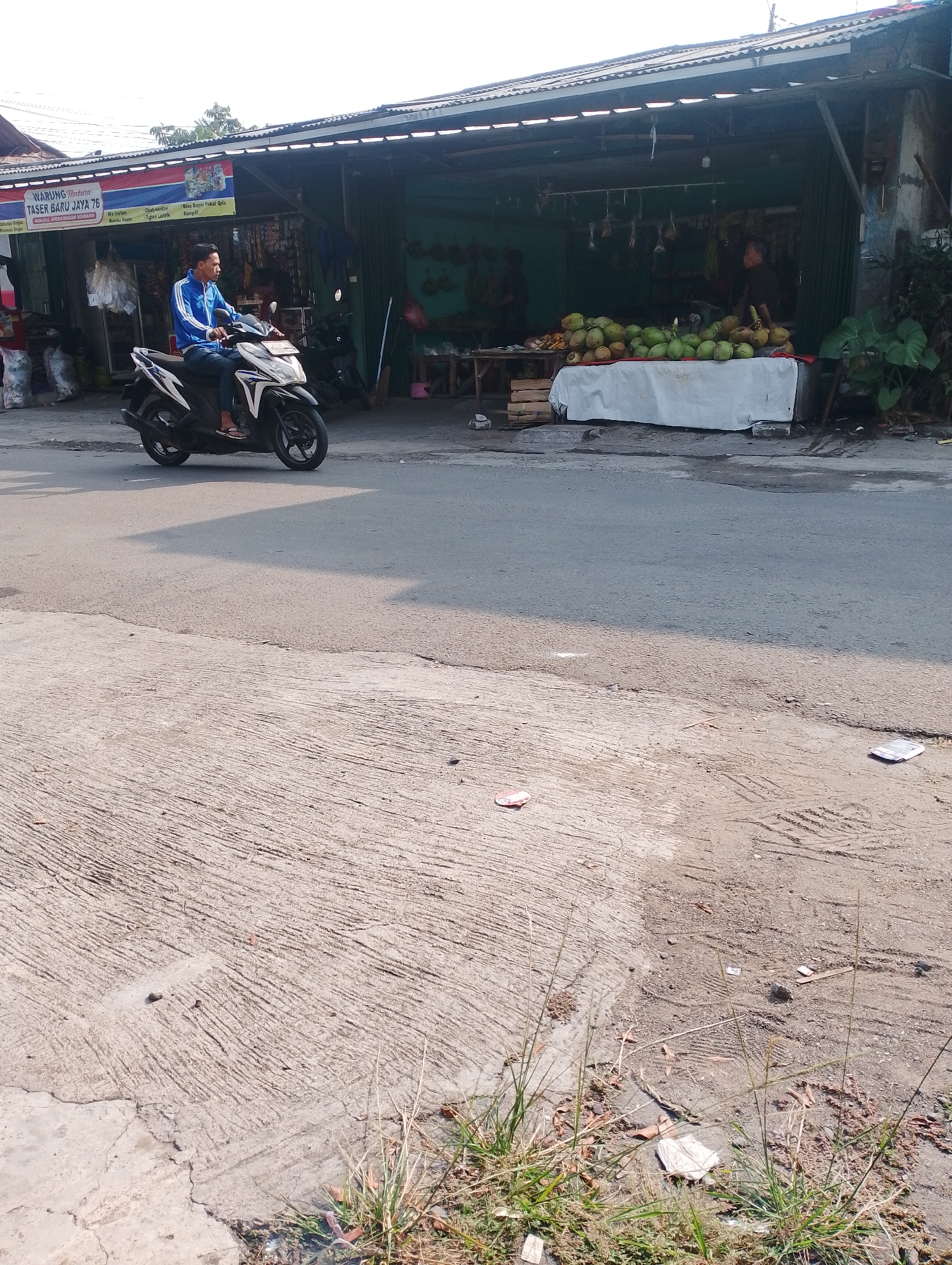 A man in a blue jacket and jeans rides a black and white scooter in front of a roadside market stall in Bogor, Indonesia. The market stall has a corrugated metal roof and displays various goods, including coconuts arranged on a table covered with a white cloth. The man is looking ahead and appears to be driving on a paved road. The environment is sunny, with shadows cast on the road and ground. The market stall is on the right side of the road, and there are also buildings and trees visible in the background. The ground in the foreground is a mixture of concrete and dirt.