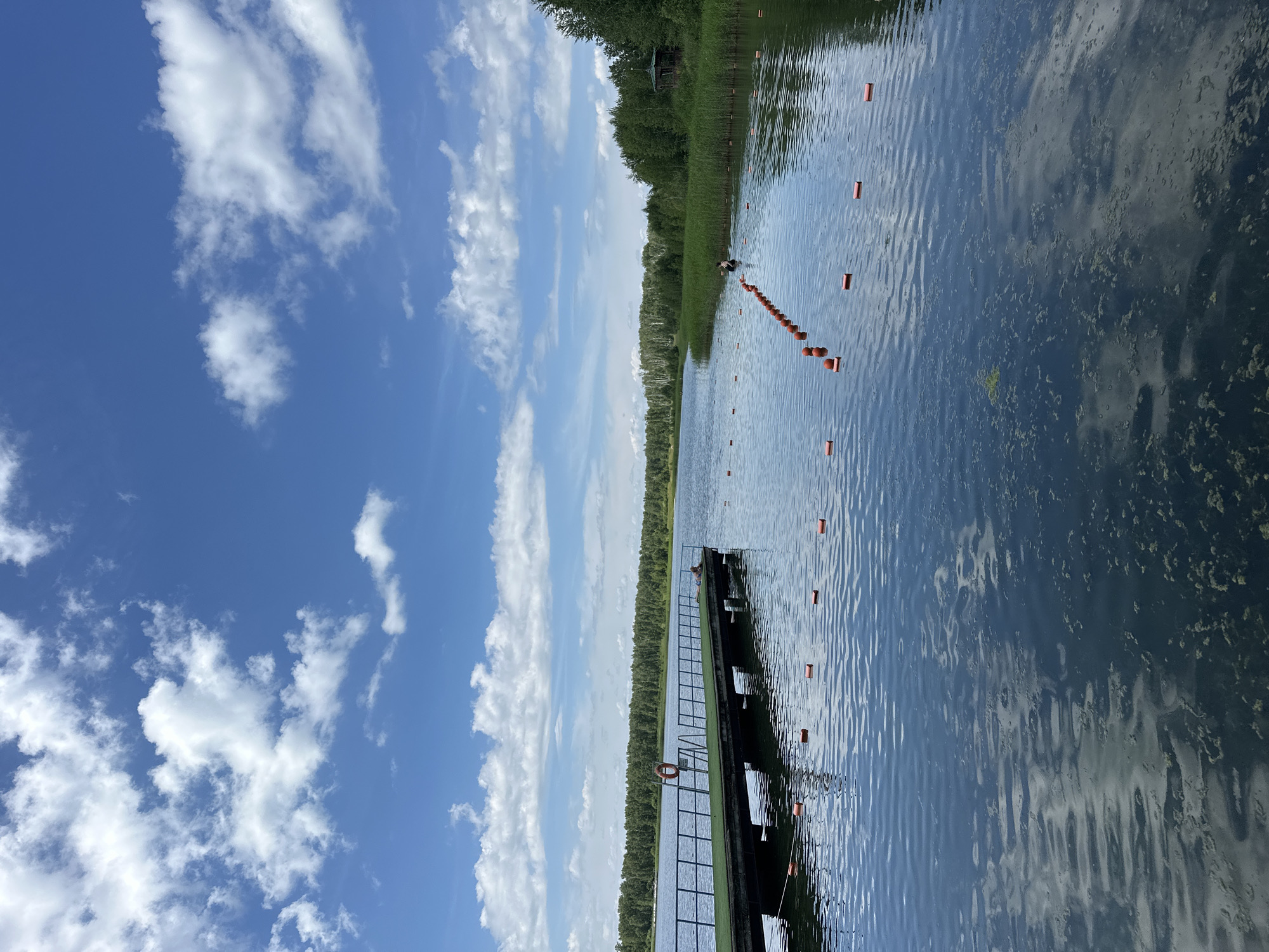 The image depicts a scenic view of the Duvankul' lake in Duvankul', Russia, on a clear day with scattered white clouds against a vibrant blue sky. In the foreground, a wooden pier extends into the tranquil water, featuring a green surface and a metal railing. A life preserver ring is attached to the railing of the pier. On the pier, a single individual is seen resting, lying down. The lake is marked with a line of orange buoys, creating a designated area within the water. In the background, a dense forest lines the opposite shore, with tall reeds growing near the water's edge. To the right of the frame, a small wooden gazebo with a roof is nestled amongst the trees. The water's surface is calm with gentle ripples, reflecting the sky and surrounding trees, and appears to have some floating aquatic vegetation in certain areas.