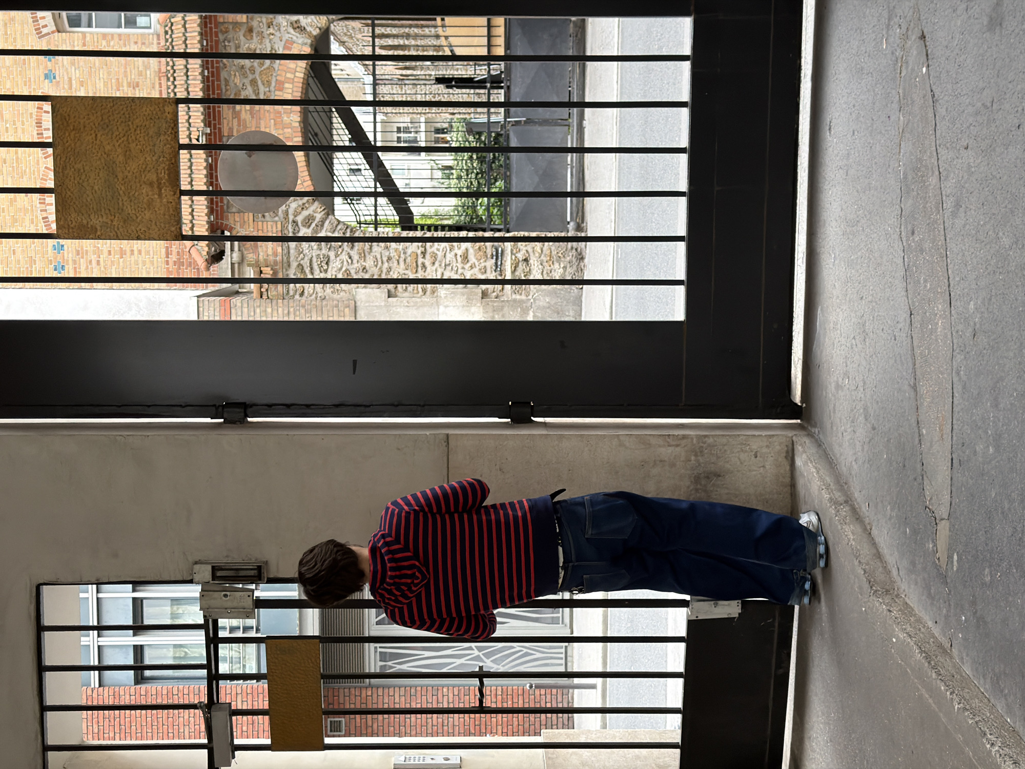 A person stands in a gray concrete passageway looking through a metal grate in Paris, France. The person wears a red and navy striped long-sleeve shirt and dark blue pants with light blue shoes. The grate divides the passageway and provides a view to the outside where a brick building and street are visible.