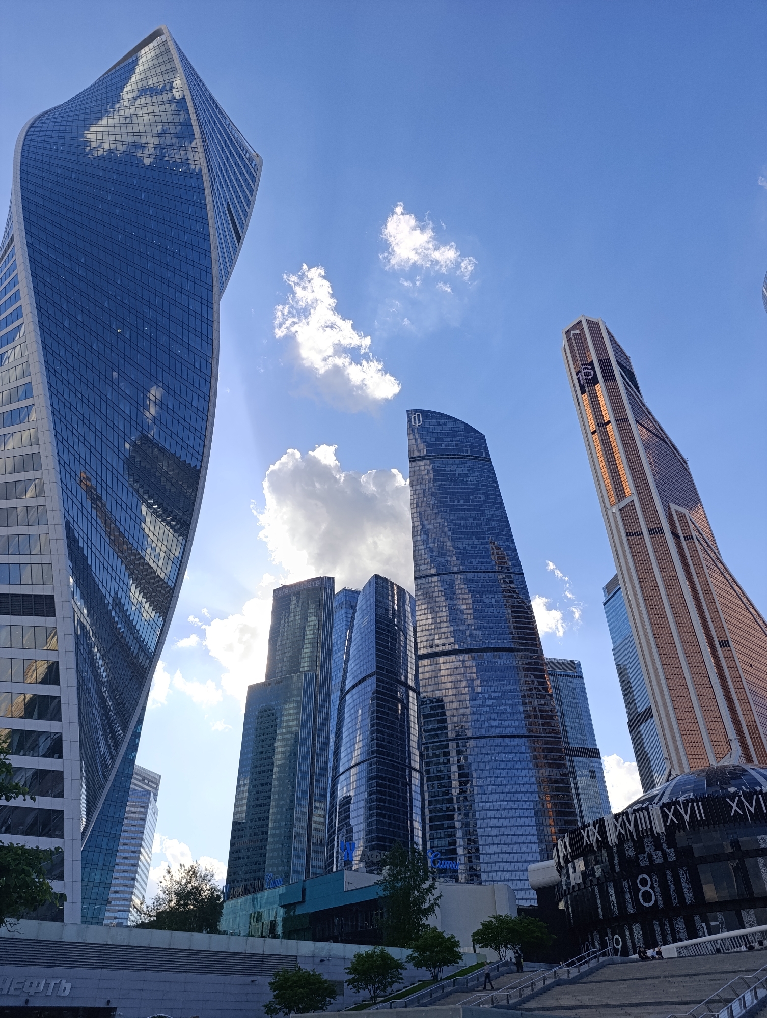The image captures a dynamic low-angle view of several modern skyscrapers in Moscow-City, Russia, under a bright blue sky with scattered white clouds. The scene is dominated by towering, glass-clad buildings reflecting the sunlight and sky.
On the far left, the distinctively twisted, blue-glass Evolution Tower rises prominently, its facade reflecting clouds. On the far right, the Mercury City Tower, distinguished by its golden/bronze-colored, tapering facade, angles upwards. Several other sleek, dark-blue glass skyscrapers stand in the middle ground, showcasing their reflective surfaces. One central building displays a logo resembling a stylized "D" or "O," while another further back bears the "ВТБ" (VTB) logo.
The time of day is sunny, with bright light creating visible sun rays emanating from behind some clouds. The ground level features various structures, including a broad set of stairs with railings, and small trees. At the bottom right, a unique domed structure with a dark, reflective surface is visible. This structure is adorned with large white Roman numerals such as "XIX," "XVIII," "XVII," and "XVI," as well as Arabic digits like "8," "1," "9," and "1-1."
A few people are scattered across the stairs and sitting on benches, enjoying the urban space. On a building at the lower left, the Russian word "НЕФТЬ" (NEFT), meaning "oil," is prominently displayed. The overall impression is one of a grand, modern architectural landscape.