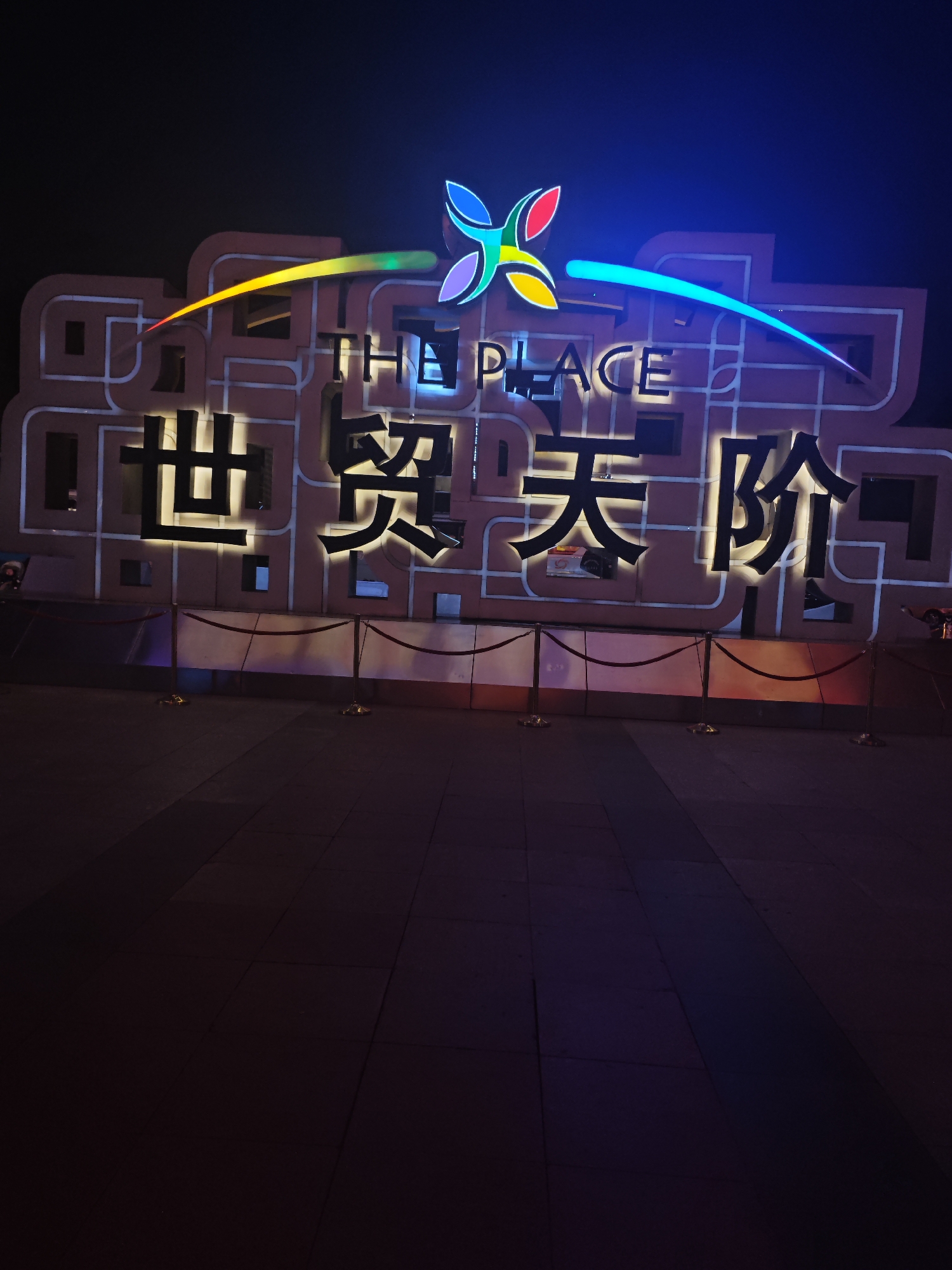 The image displays a brightly illuminated outdoor sign at night, located in what appears to be a commercial or entertainment area within Chaoyang District, China. The scene is dominated by the large, glowing signage, with the surrounding environment largely dark.
The primary subject is a massive sign structure that features both English and Chinese text, along with a prominent, colorful logo at its apex. The logo resembles a stylized, multi-colored butterfly or a four-petal flower, with segments in blue, red, yellow, and green. Two long, illuminated arcs extend horizontally from the logo, with one showing a gradient from yellow to green on the left, and the other a solid blue hue on the right.
Below the logo, the English words "THE PLACE" are displayed in large, luminous white letters. Underneath the English, larger Chinese characters are visible, also illuminated with a warm, soft white light. These characters read "世贸天阶" (Shì mào tiān jiē), which translates to "The Place" or "World Trade Heaven Street," confirming the location as the well-known shopping and entertainment complex in Beijing. The background structure of the sign appears to be a reddish-brown panel with geometric patterns and embedded lighting.
In front of the sign, several decorative barriers with red ropes and golden stanchions are set up, likely to manage crowd flow or mark an entrance. The ground in the foreground is a dark, paved surface, reflecting subtle hints of the colorful lights from above. There are no people visible in the image, emphasizing the architectural and lighting design of the landmark. The time of day is clearly night, as indicated by the dark sky and the sign's illumination.