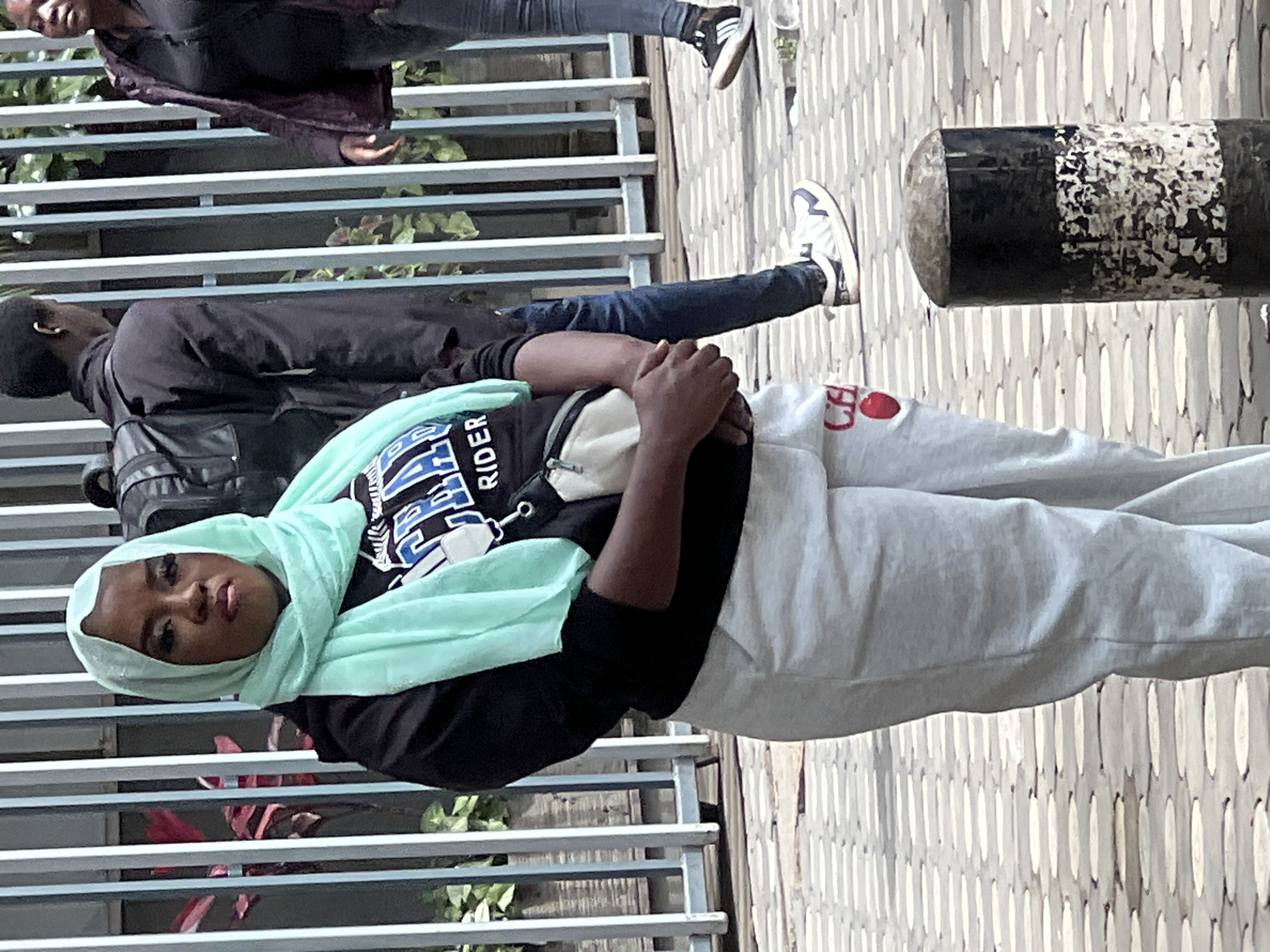 The image shows a woman wearing a light blue hijab and a black shirt with a "RIDER" design. She's wearing light grey sweatpants with a red design. She's positioned in front of a grey metal railing. Another person is standing behind her, and a third individual is partially visible at the top of the frame. The ground appears to be a patterned walkway. Based on the architectural style and the people, it can be inferred that the photo was taken in an urban environment, potentially Nairobi, Kenya.