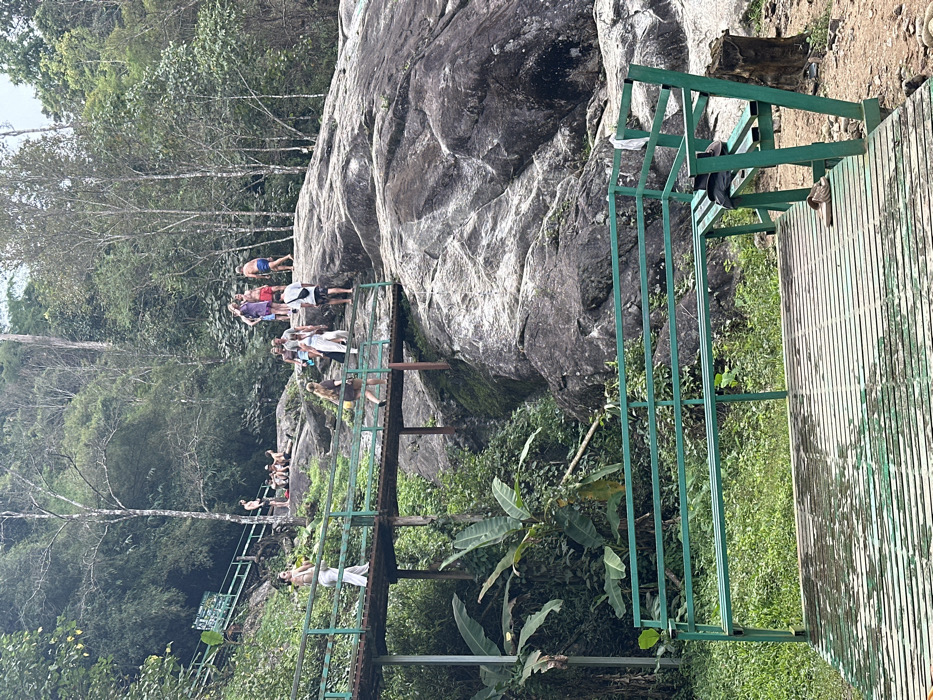 A lush, vibrant outdoor scene features a rocky formation adjacent to a series of green metal railings and a wooden walkway. A group of people stand atop the rocks and walk along a pathway extending from the side. Dense foliage surrounds the scene, with trees and banana plants adding depth and a tropical feel. The shot captures a natural setting popular for outdoor activities, likely near a waterfall or natural pool.