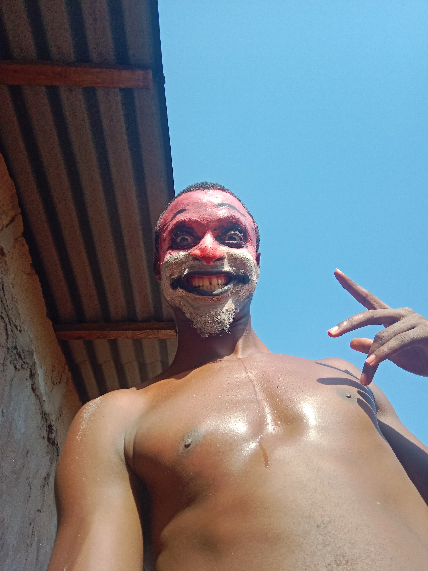 A shirtless man with red and white face paint stands outdoors in Nkpor, Nigeria. The sky is blue. He is smiling and giving a peace sign. He is positioned between a corrugated metal roof and a dirt wall.