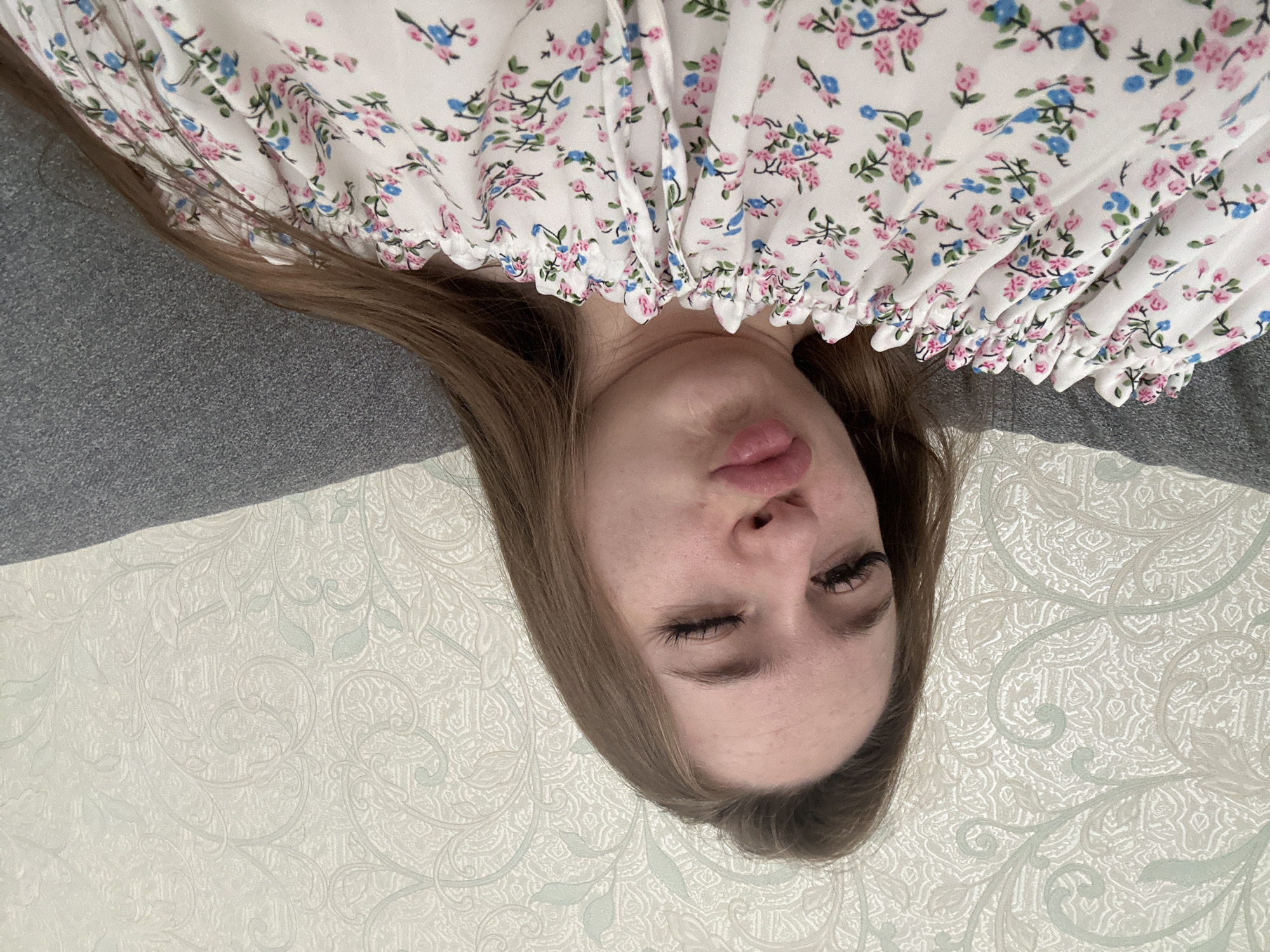 The image displays an upside-down, close-up view of a young woman lying down indoors. Her head is positioned towards the bottom of the frame, framed by her light brown hair. Her eyes are mostly closed, and her lips are slightly puckered, suggesting a playful expression or a gentle pout. Long eyelashes are visible.
She is wearing a white garment, likely a top or dress, adorned with a delicate floral pattern composed of small pink and blue flowers with green leaves. The fabric around her neck area is gathered or ruffled, creating soft folds.
The background consists of two different textures. To one side of her head, there is a solid, medium-grey fabric, possibly part of a sofa or bedding. On the other side and extending beneath her, there is a lighter-colored surface with an intricate, embossed or jacquard-like pattern featuring ornate, swirling motifs, resembling wallpaper or patterned bedding.
The scene conveys a relaxed, intimate indoor setting, likely a home environment. No text is visible in the image.