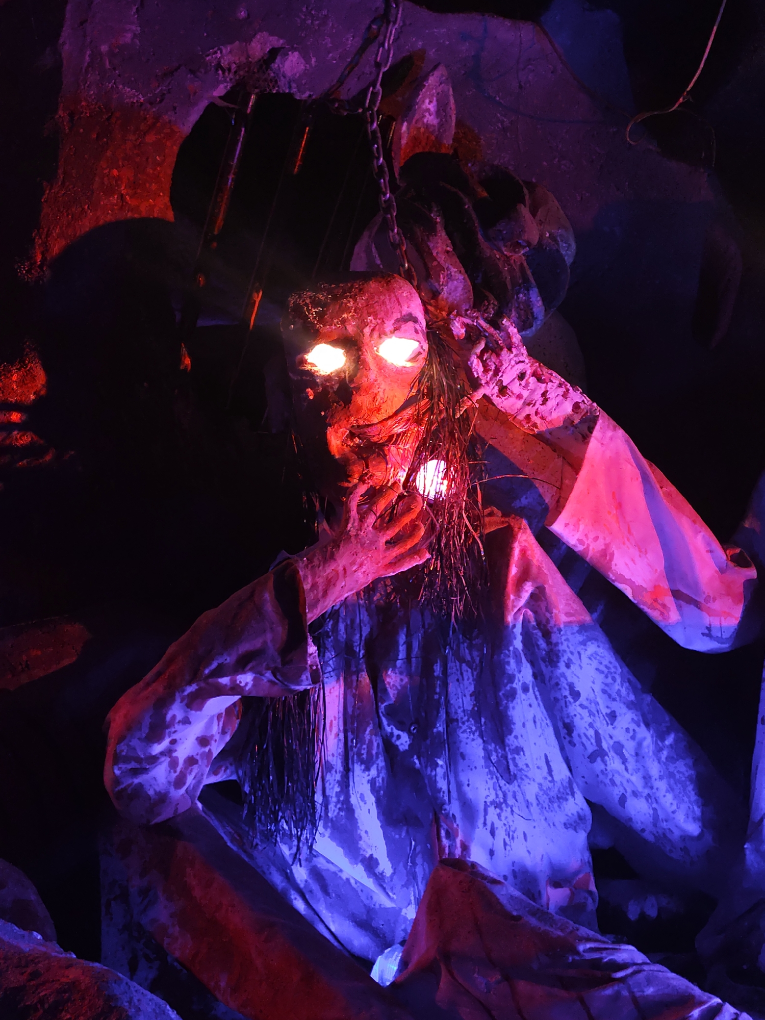 The image depicts a gruesome, horror-themed scene, likely part of a haunted house attraction. A humanoid figure, possibly a mannequin or a prop, is the focal point. The figure is suspended by chains within a dark, cave-like environment, its eyes illuminated with an eerie, bright glow. The figure is dressed in a tattered, white garment stained with what appears to be blood. Long, dark strands of hair hang down, further obscuring the figure's face. The scene is lit with a combination of red and blue lighting, creating an unsettling atmosphere. The surrounding environment consists of rough, rocky surfaces suggesting a subterranean setting.