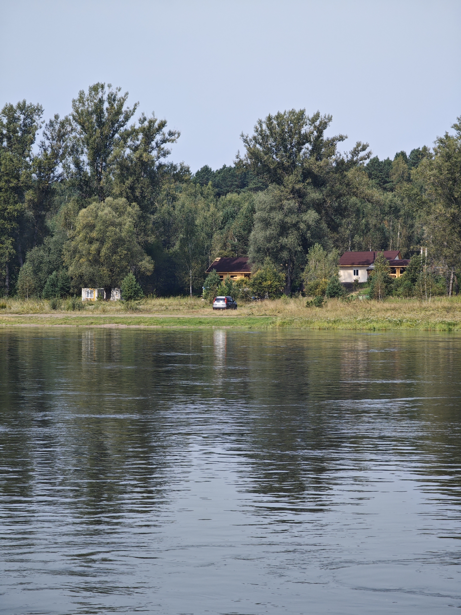 The image depicts a serene river scene with a densely wooded shoreline. In the foreground, the calm river reflects the sky and the trees on the opposite bank. The middle ground shows a grassy bank with several buildings and a parked car. A small, weathered building with a yellow facade and a white-framed window is situated on the left. Adjacent to it, a taller, modern house with a brown roof and light-colored walls stands on the right, with a wooden porch visible. A silver SUV is parked on the grass in front of the house. The background is dominated by a thick forest of various trees, suggesting a natural, possibly rural, setting. The sky above is clear and pale blue. The location is identified as the city of Kononovo, Russia.