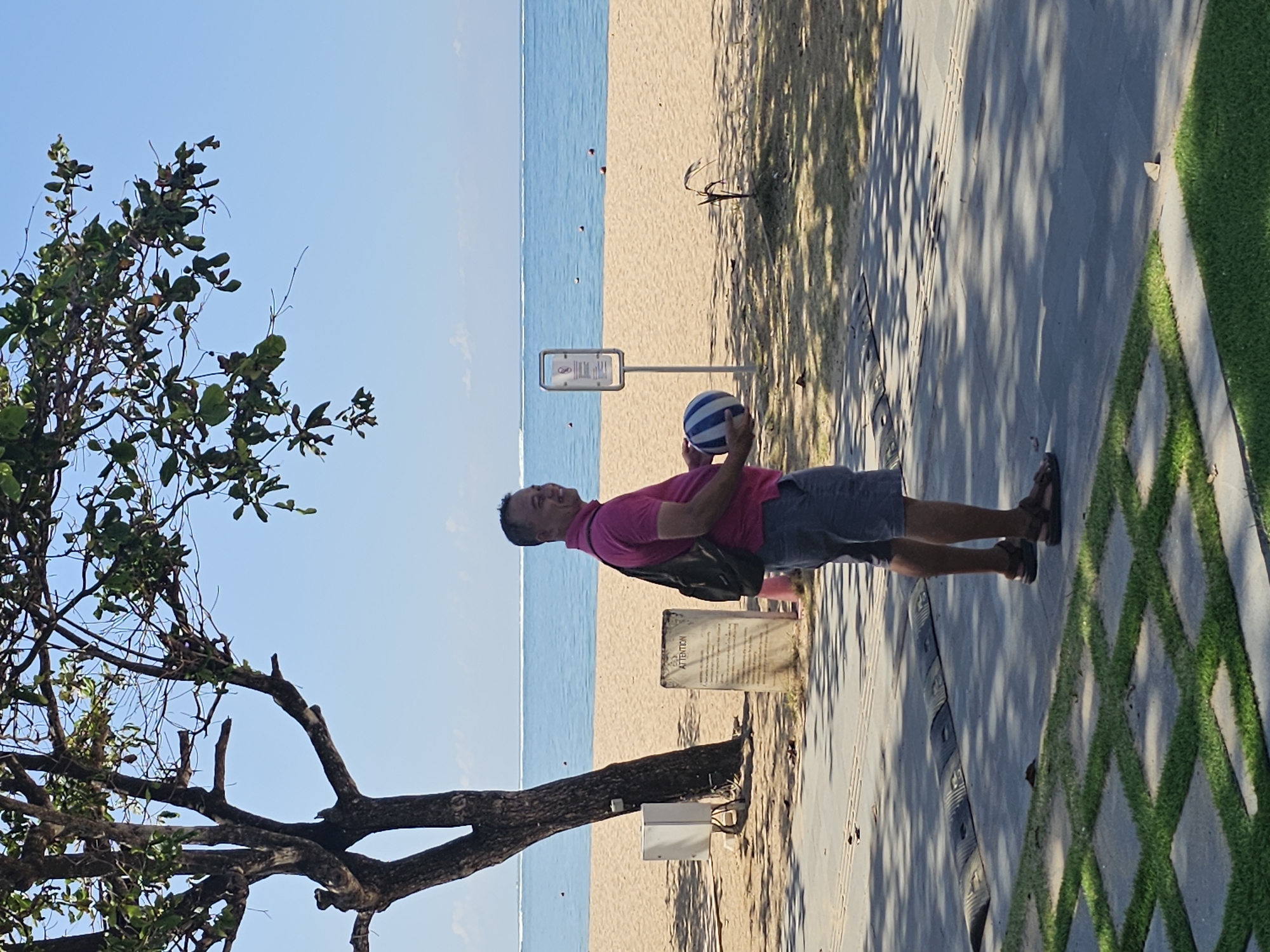 A man stands on a paved walkway next to a beach in Denpasar, Indonesia. He is wearing a pink polo shirt, gray shorts, and brown sandals. The man is holding a blue and white striped ball and has a backpack over his right shoulder. Behind him, a sandy beach meets the blue ocean, with a clear sky overhead. A tree with green leaves is to the left of the man, casting shadows on the walkway. A sign with the word "ATTENTION" is visible on the beach. The walkway has textured surfaces and a speed bump. Green grass is planted in a grid pattern in the foreground.