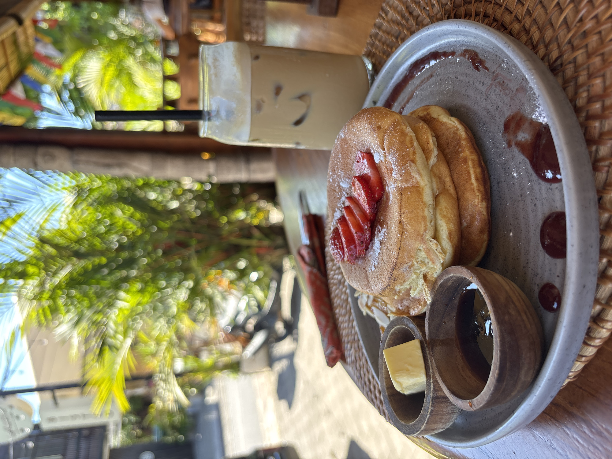 A breakfast scene is set outdoors in Pecatu, Indonesia, featuring pancakes and an iced coffee drink. The photograph is taken from an oblique, slightly downward angle.
In the foreground, a stack of three pancakes dusted with powdered sugar and topped with sliced strawberries is the focal point. They rest on a gray, earthenware plate adorned with swirls and droplets of red jam or sauce. Two small, wooden ramekins—one containing a pat of butter and the other filled with a dark amber liquid, likely syrup or honey—are positioned next to the pancakes. The plate sits on a round, woven placemat of natural fibers.
Behind the plate, an iced coffee drink in a tall, square glass is visible, containing ice cubes and a straw. The background blurs into an outdoor setting. Out-of-focus foliage, including palm trees, provides a backdrop, suggesting a tropical location. Hints of building facades are vaguely discernible, with dappled sunlight filtering through the leaves.