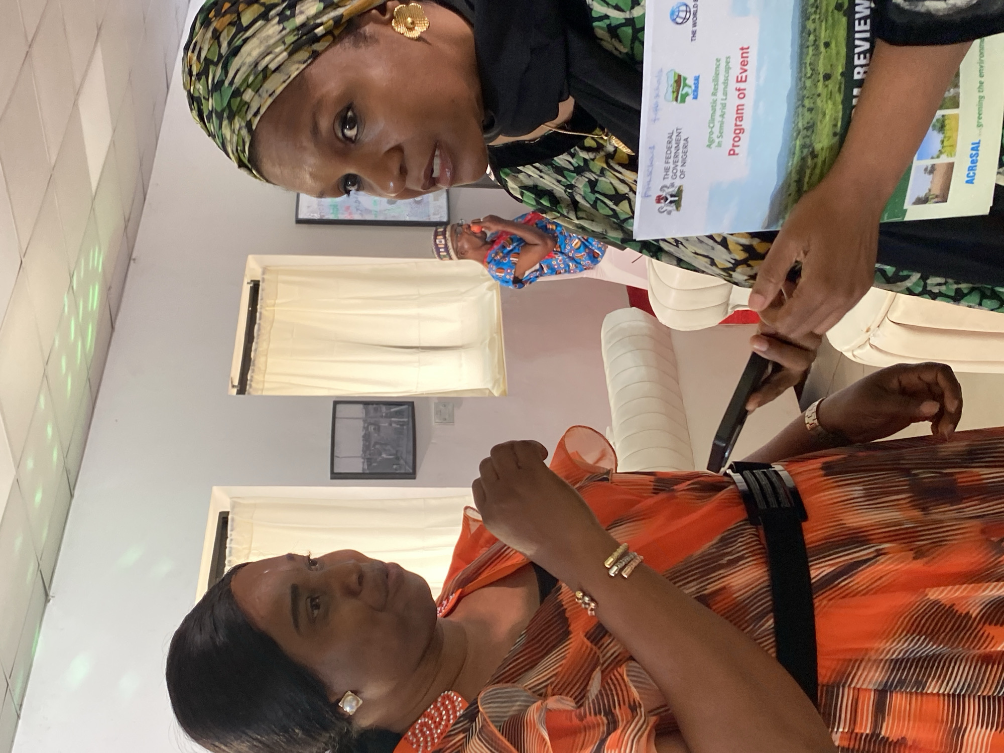 Two women are in conversation, possibly at an event. The woman on the right, wearing a patterned headscarf and gold earrings, holds a program titled "Agro-Climatic Resilience in Semi-Arid Landscapes: Program of Event" with logos for the Federal Government of Nigeria and the World Bank. The other woman, in an orange dress with ruffle details and a black belt, looks up at her. She is wearing earrings and bracelets. A phone is visible in the woman's hand on the right. Two framed pictures are affixed to the wall behind the women. Based on the program documentation, the setting is possibly in the city of Kwarau, Nigeria, given the Federal Government of Nigeria and landscape imagery.