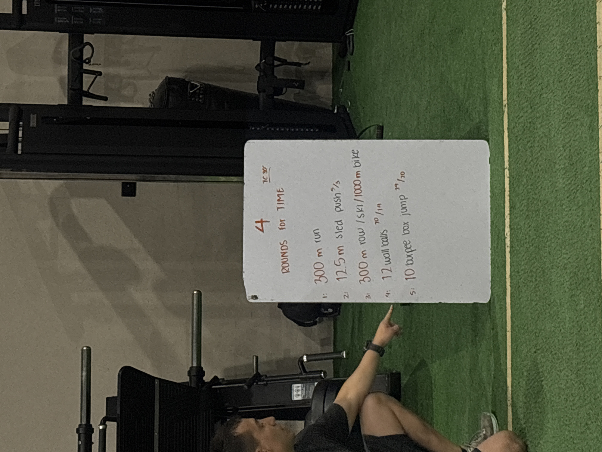 The image displays an indoor gym environment where a person is pointing at a whiteboard detailing a workout routine. The individual, visible from the chest down, is seated on a green artificial turf floor marked with white lines, wearing dark athletic shorts and a watch on their left wrist. Their right index finger is extended, drawing attention to the text on the whiteboard.
The whiteboard prominently features a workout titled "4 ROUNDS for TIME," with a "TC 30'" (Time Cap 30 minutes) noted at the top. The regimen consists of five exercises:
1. "300 m run"
2. "12.5m sled push 2/3" (likely referring to a specific sled configuration or weight)
3. "300 m row / ski / 1000 m bike" (providing options for cardiovascular exercise)
4. "12 wall balls 20/14" (suggesting medicine ball weights of 20 lbs for men and 14 lbs for women)
5. "10 burpee box jump 24/20" (indicating box heights of 24 inches for men and 20 inches for women)
In the background, dark gym equipment, possibly a weight rack or a leg press machine, and a dark bag are discernible. The setting is clearly an indoor fitness facility, illuminated by artificial lighting, and the scene implies a workout session is being reviewed or explained.