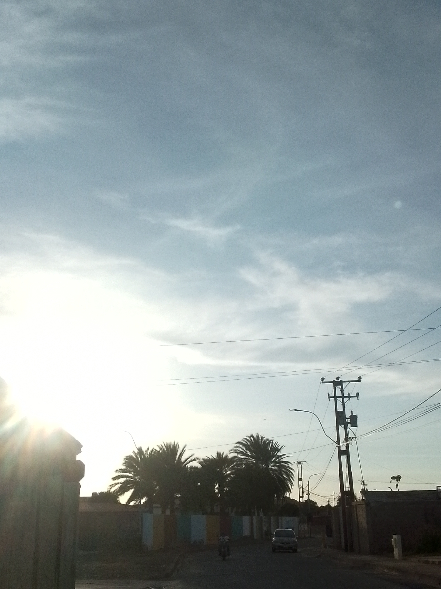 The image presents an outdoor street scene at either sunrise or sunset, dominated by a brightly lit sky and silhouetted foreground elements. The upper half of the frame features a light blue sky with scattered, wispy white clouds. The sun, positioned intensely on the far left, creates a strong glare and lens flare, backlighting much of the scene.
In the foreground on the left, a dark, partially visible structure, possibly a gate or wall, is heavily shadowed by the sun. In the mid-ground, several tall palm trees are distinctly silhouetted against the bright horizon. Behind them, parts of buildings with faintly discernible multi-colored vertical stripes (blue, white, yellow, brown) are visible.
On the right side of the image, a tall utility pole with numerous electrical lines, a transformer, and an attached street lamp stands prominently, also appearing as a dark silhouette. Multiple power lines crisscross horizontally across the sky.
The asphalt road in the lower portion of the image appears somewhat wet in areas, reflecting some light. Two vehicles are visible: a dark-colored car driving away from the viewer in the mid-ground, and a person on a motorcycle further down the road on the left.
The strong sunlight, palm trees, and general urban landscape suggest a warm climate, consistent with the provided location context of Juan Griego, Venezuela. No discernible text is visible in the image.