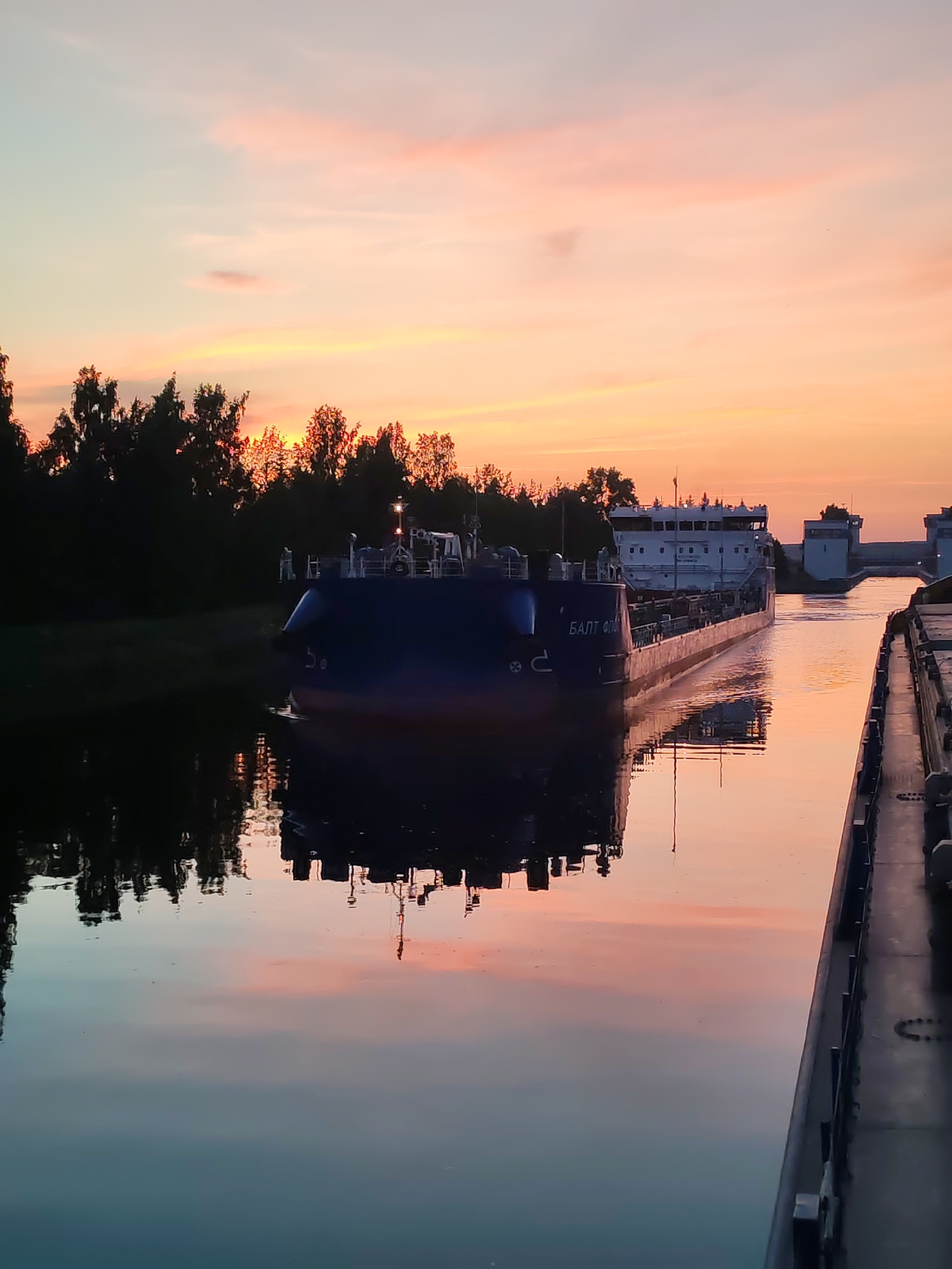 The image shows a sunset over a canal in Vytegra, Russia. A large barge, identified by the Cyrillic text "БАЛТ ФЛОТ" on its side, is navigating the canal. The barge's reflection is visible in the calm water, which is illuminated by the warm hues of the sunset sky. Silhouetted trees line the canal's edge, and further in the distance, structures resembling canal locks can be seen. The sky transitions from a pale blue at the top to soft pinks and oranges near the horizon. There are no people clearly visible in the image.