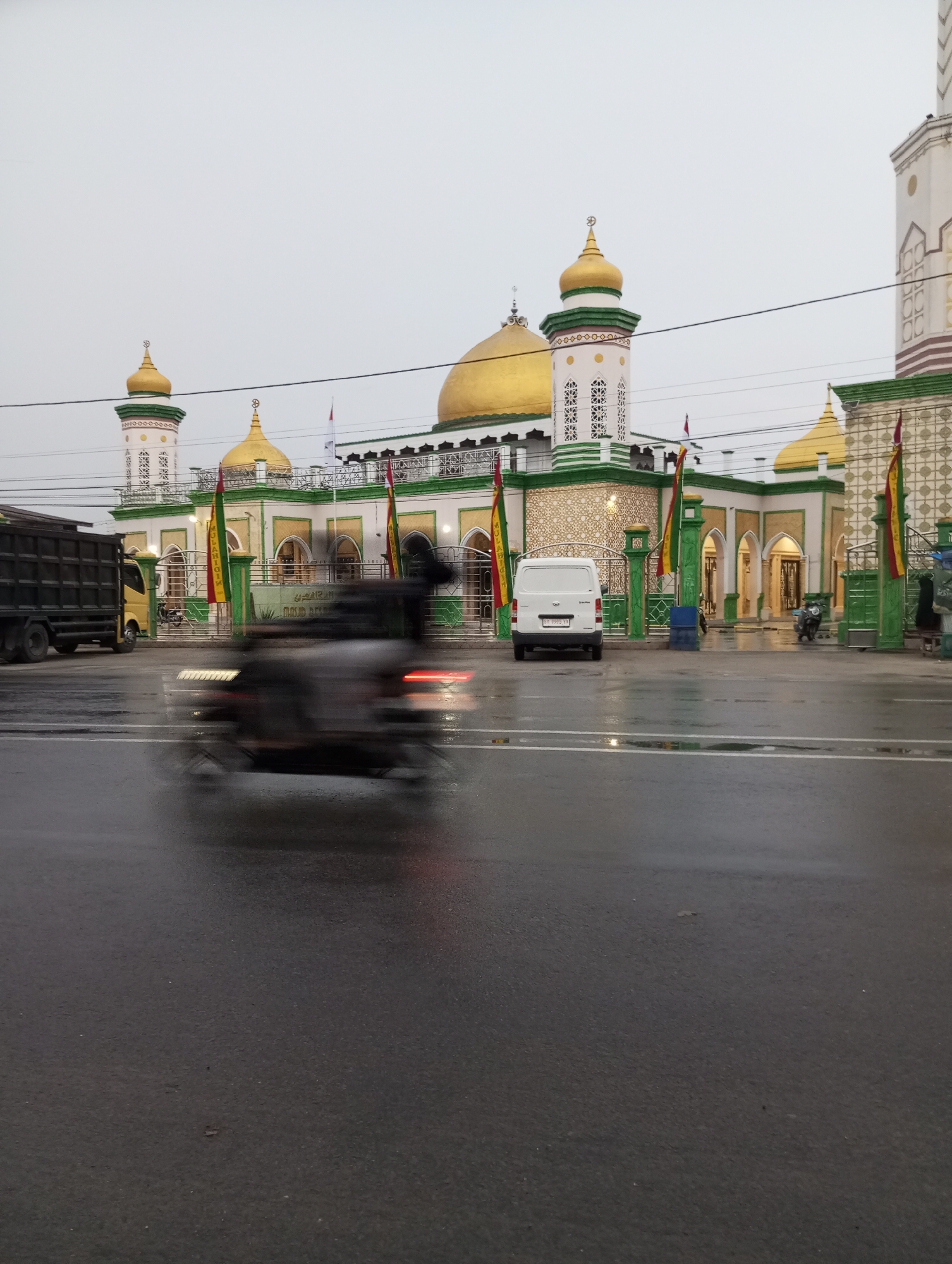 The image captures a street scene in Air Molek, Indonesia, with a mosque as the focal point. The mosque is characterized by its prominent golden domes and green accents on its white structure. Two minarets, also topped with golden domes, flank the main building. The mosque is decorated with flags and intricate patterns on its facade. In the foreground, a wet asphalt road reflects the overcast sky. A blurred motorcycle, indicating motion, is passing by. To the left, a dark-colored truck is parked, and further to the right, a white van is visible. The environment appears to be a public area with some greenery and fencing around the mosque's entrance. The sky is overcast, suggesting a cloudy or rainy day.