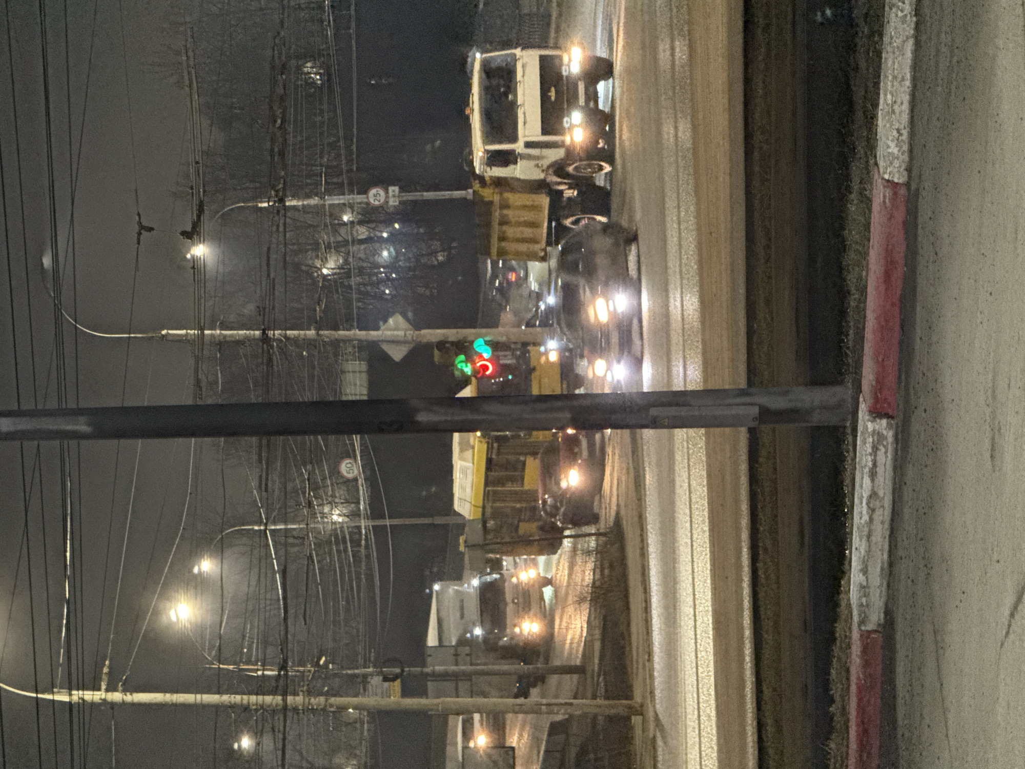 The image shows a street scene at night in Izhevsk, Russia. There are several vehicles on the road, including a large white truck, a yellow bus, and several cars with their headlights on. The traffic light is green. Streetlights are visible along the road, and utility poles with many wires are on the left side of the street. A concrete barrier with red and white markings is in the foreground on the right. The road appears wet. A road sign showing speed limit 50 is seen along the road.