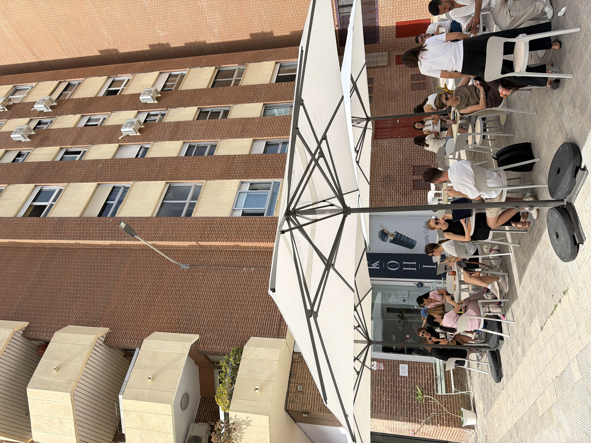 In an outdoor seating area in front of a building, several people are seated at tables under large white umbrellas. The building facade is made of red brick with cream-colored sections and balconies. Air conditioning units are visible on the building. On the left, a small tree is planted in a white pot. People are interacting, some looking at phones, others talking or looking at laptops. One woman is standing. The ground is paved with square tiles. A sign for "KOHL Specialty Coffee" is visible on the building entrance.