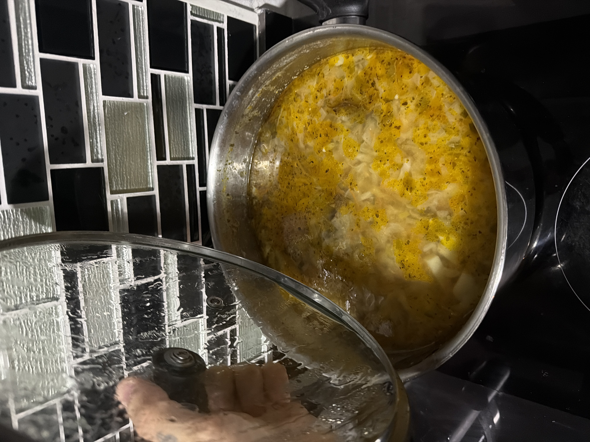 A person's hand is lifting the lid off a pot of soup on a stovetop in a kitchen. The soup is a vibrant yellow with visible herbs and chunks of vegetables. The stovetop appears to be an induction cooktop. Behind the pot, a backsplash of black and glass mosaic tiles is visible. The scene does not provide any information that would allow for its location to be identified as Bangkok, Thailand.