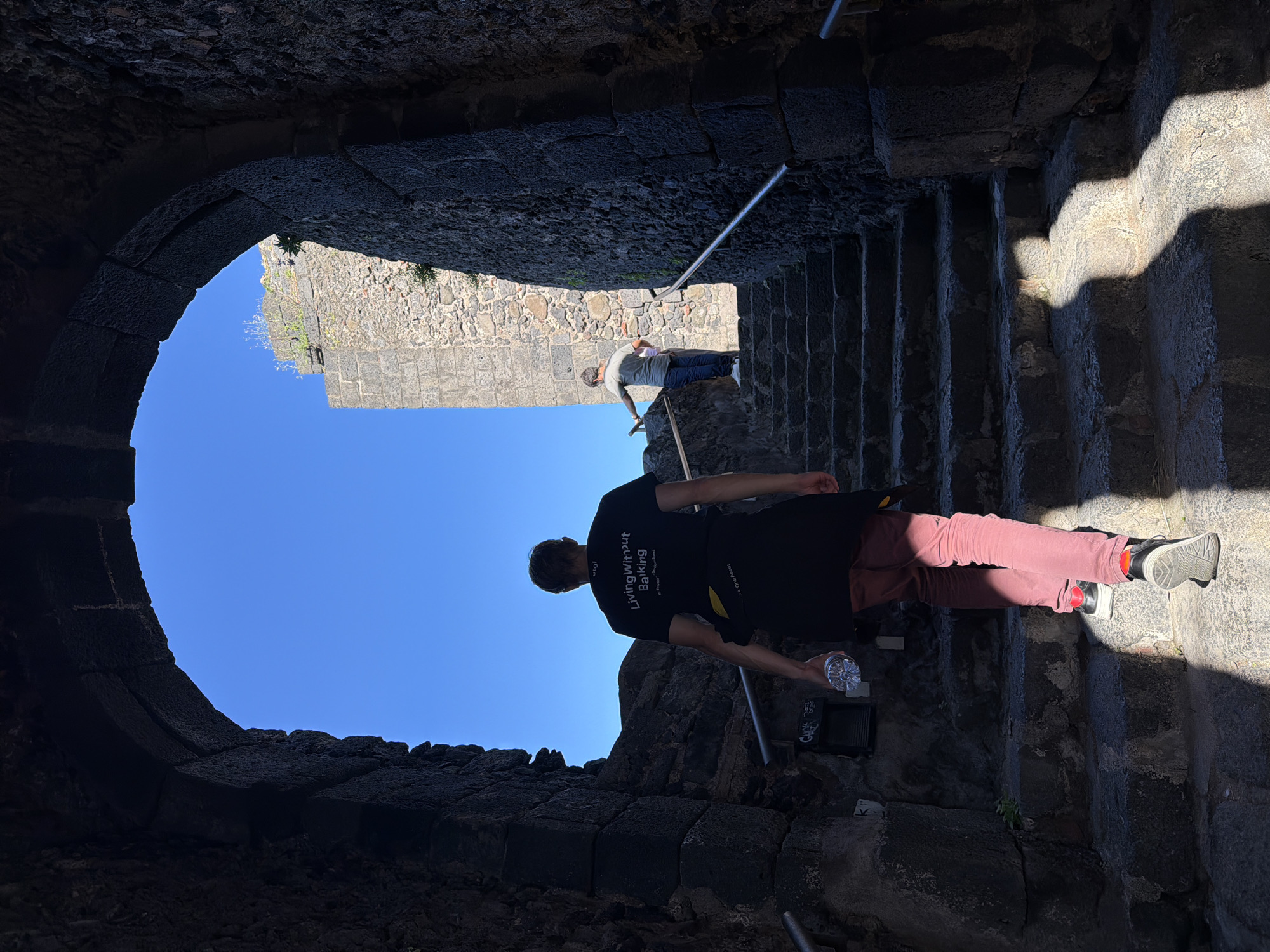 The image captures a scene within Aci Castello, Italy, showcasing a person descending stone steps inside the castle. The individual wears a black t-shirt with white writing on the back, dark pants, and sneakers with red accents; they carry a water bottle. The stairs are stone and lead down from an arched opening, through which a blue sky and parts of the castle walls are visible. A second person is visible further up the stairs. The environment is characterized by the rough texture of the stone walls and steps.