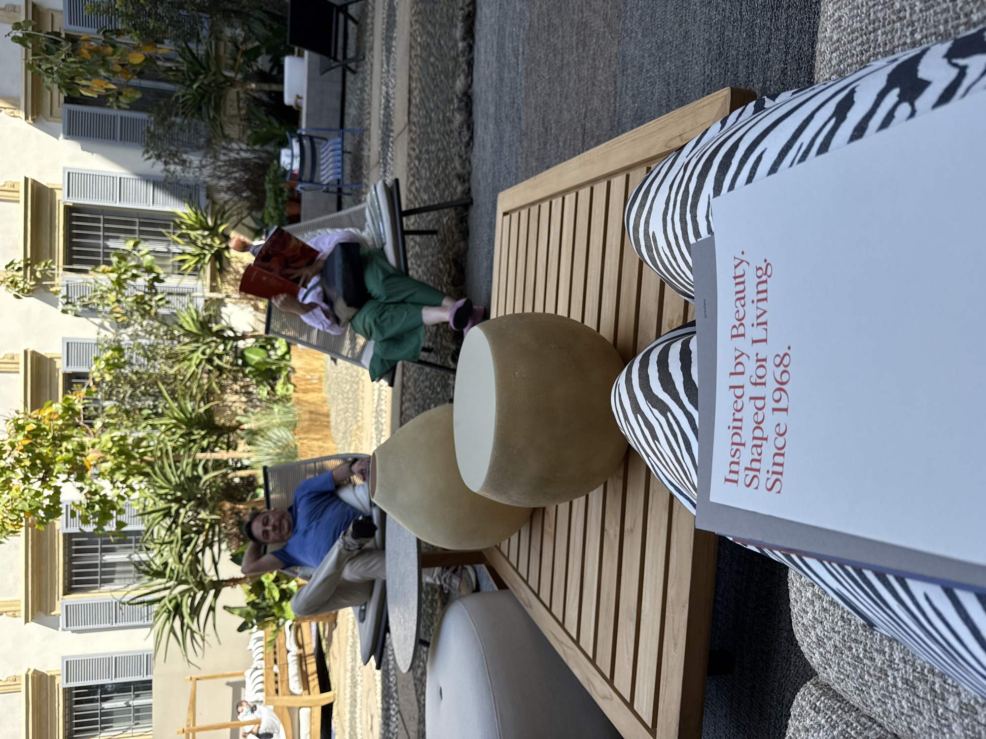 The image shows an outdoor seating area in what appears to be Milan, Italy, judging by the architecture. Two people are visible: one man is lounging in a chair, wearing a blue shirt and beige pants, while a woman is sitting in another chair reading a book, dressed in a white shirt, green pants, and pink slippers. The seating area includes modern furniture, such as rounded side tables, zebra-print patterned chairs, and a wooden-slatted table. A large book is visible on the zebra-print chair. The backdrop includes a light-colored building with blue-grey shutters, abundant greenery, and a pebbled ground surface.