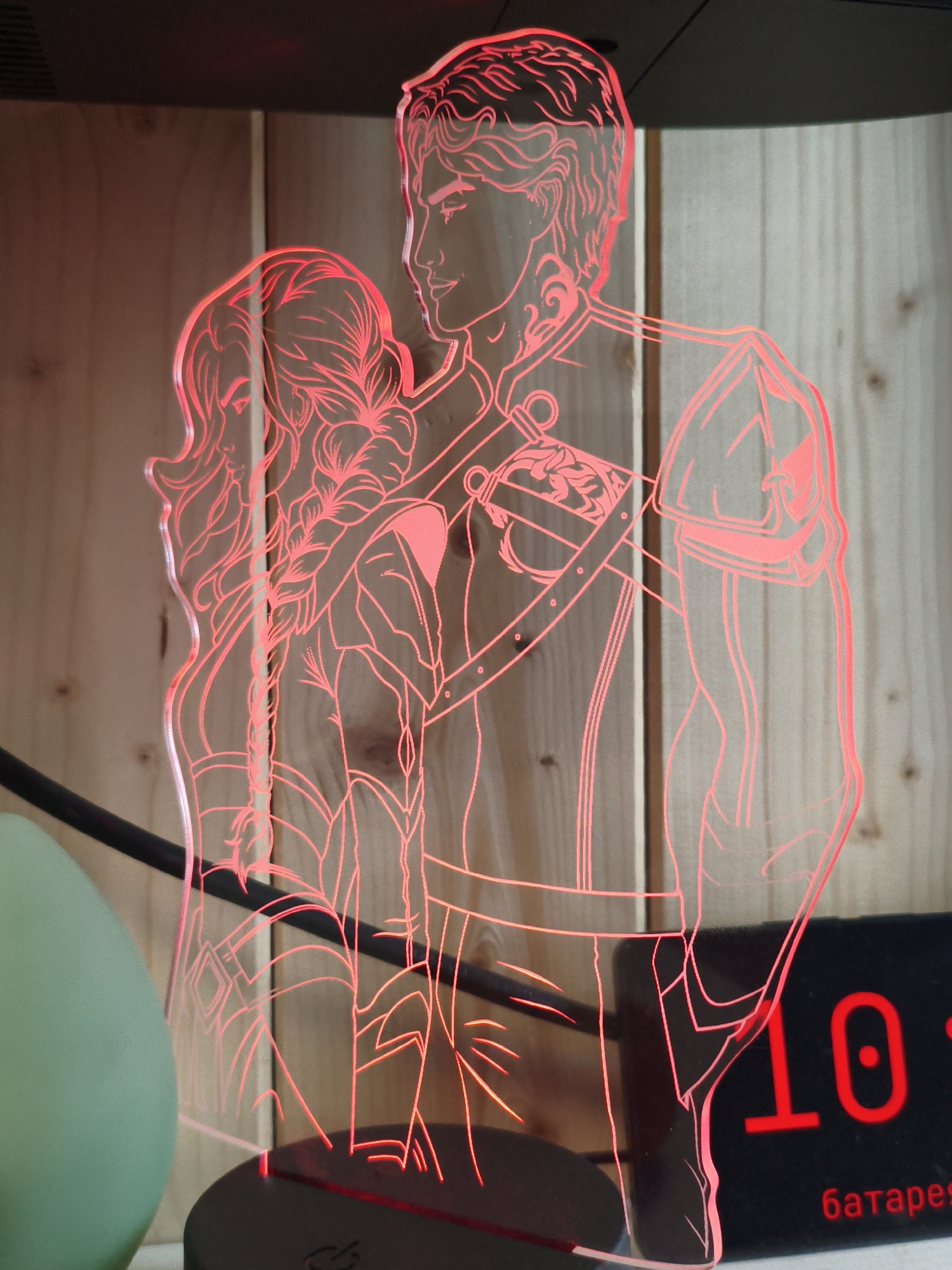The image shows a red-lit acrylic lamp featuring a line art depiction of a couple. The man is on the right, facing left in profile, with his arm around the woman. The woman is on the left, facing right in profile, with her head resting on the man's shoulder. Both figures have stylized hair and wear clothing that appears to be historical or fantasy-inspired. The lamp is illuminated by a red light, creating a glowing effect on the acrylic.
The lamp is positioned on a dark base, which is not fully visible. Behind the lamp, a wooden background with vertical planks is visible. To the left of the lamp, a portion of a large, light green, spherical object is present. To the right of the lamp, a black digital clock with red illuminated numbers is partially visible. The clock displays "10:" and below that, the Russian word "батарея" (battery).
The scene is indoors, and the lighting suggests it is likely nighttime or in a dimly lit room. The overall impression is that of a decorative item, possibly a night light or desk lamp.
There is no indication in the image that this scene is located in Vladivostok, Russia. The presence of the Russian word on the clock is the only element that could suggest a Russian context, but this does not confirm the specific city.