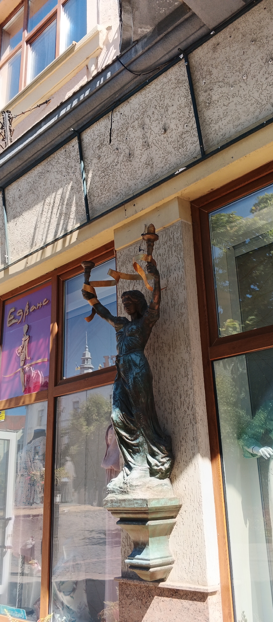 A bronze statue of a woman holding two torches is mounted on the exterior wall of a building in Mukachevo, Ukraine. The statue is dark green and appears weathered. The woman is depicted wearing a long, flowing dress and has her arms raised, holding the torches aloft. A yellow ribbon is wrapped around the torches and her arms. The building has large windows, some of which reflect the sky and trees. One window displays a purple sign with an image of a stylized human figure. The statue is mounted on a decorative pedestal, which is affixed to the building's textured, light-colored wall. The overall scene suggests an urban environment with architectural elements and artistic embellishments.