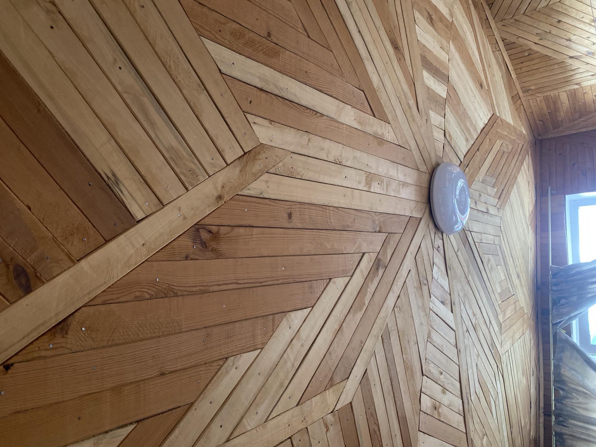 The image shows a wooden ceiling with a geometric pattern formed by planks of varying widths and shades of brown. The planks are arranged in a radiating design, converging towards a central circular light fixture. Some planks are oriented diagonally, creating a dynamic and textured appearance. The wood grain is visible, and small metal nails are apparent, securing the planks. In the lower portion of the image, a section of a window with curtains is visible, suggesting an interior space. The overall impression is one of rustic craftsmanship. The provided location of Krivodanovka, Russia, is noted.