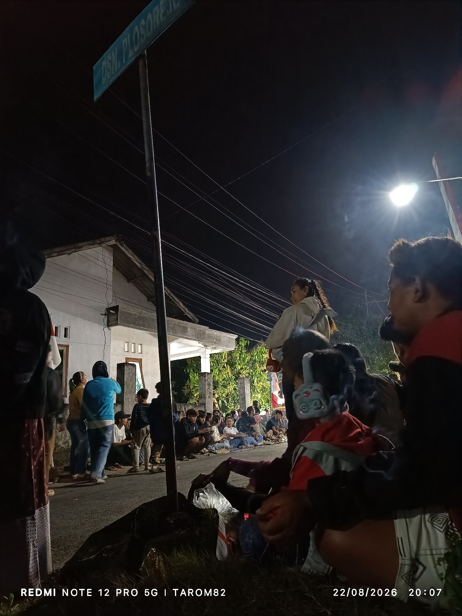 A low-angle, night-time photograph captures a street scene in Mundusewu, Indonesia. A street sign, partially legible as "DSN. PLOSORE," is mounted on a pole in the foreground. A group of people are gathered, some sitting on the ground, others standing. A house with white walls and multiple windows is visible in the background. The lighting appears to be from a street lamp and possibly the moon, casting shadows and creating a dim atmosphere. The image is watermarked with "REDMI NOTE 12 PRO 5G | TAROM82" and the date "22/08/2026" with the time "20:07".