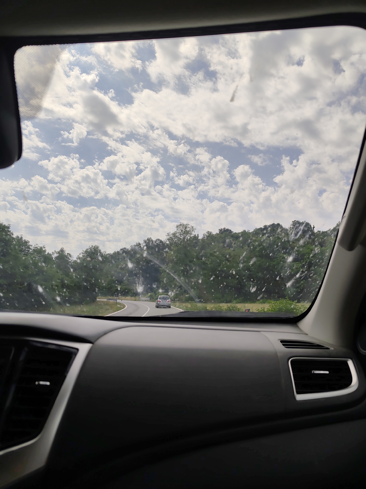 The image shows a view from inside a car, looking out through the windshield. The scene outside is a winding road going through a lush, green forest. A silver car is visible driving ahead on the road, with a cloudy sky overhead. The time of day appears to be daytime, likely late morning or early afternoon, given the bright sky and clear visibility. The weather seems to be partly cloudy, with a mix of sun and clouds in the sky. There are some water spots or debris on the windshield, which partially obscure the view. The interior of the car is visible in the foreground, showing the dashboard and air vents, which are dark gray and black. The overall mood is one of travel and forward motion, perhaps on a scenic drive. No people are clearly visible. There is no visible text in the image.