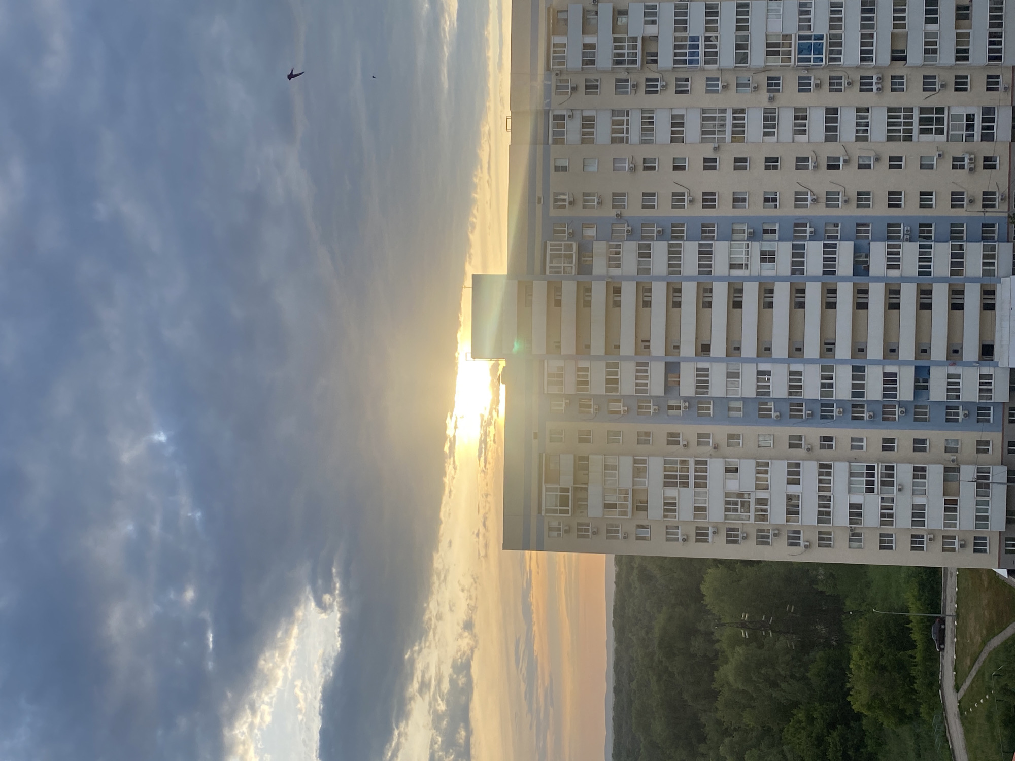 This media file depicts an urban landscape at sunset. The primary subjects are a tall, multi-story apartment building and a dramatic sky filled with clouds. The building is characterized by its repetitive rows of windows and a facade that alternates between cream and pale blue sections, with visible air conditioning units on many floors.
The scene is illuminated by the warm glow of the setting sun, which is peeking through the clouds, creating streaks of light and casting long shadows. The sky itself is a mix of dark, brooding storm clouds and lighter, more illuminated areas where the sun's rays break through, suggesting dynamic weather conditions. A small, dark silhouette, likely a bird, is visible in the upper left portion of the sky.
Below the apartment building, a dense line of green trees borders the urban area, transitioning into a more distant, hazy horizon. A paved path with some white markings and a solitary car are visible in the lower right, hinting at a suburban or edge-of-city location.
The overall mood is one of transition and dramatic natural beauty contrasting with the man-made structure. The time of day is clearly sunset, indicated by the low angle of the sun and the warm, golden light. The location is identified as Novokuybyshevsk, Russia, though no specific landmarks are clearly discernible from the image itself. There is no visible text in the image.