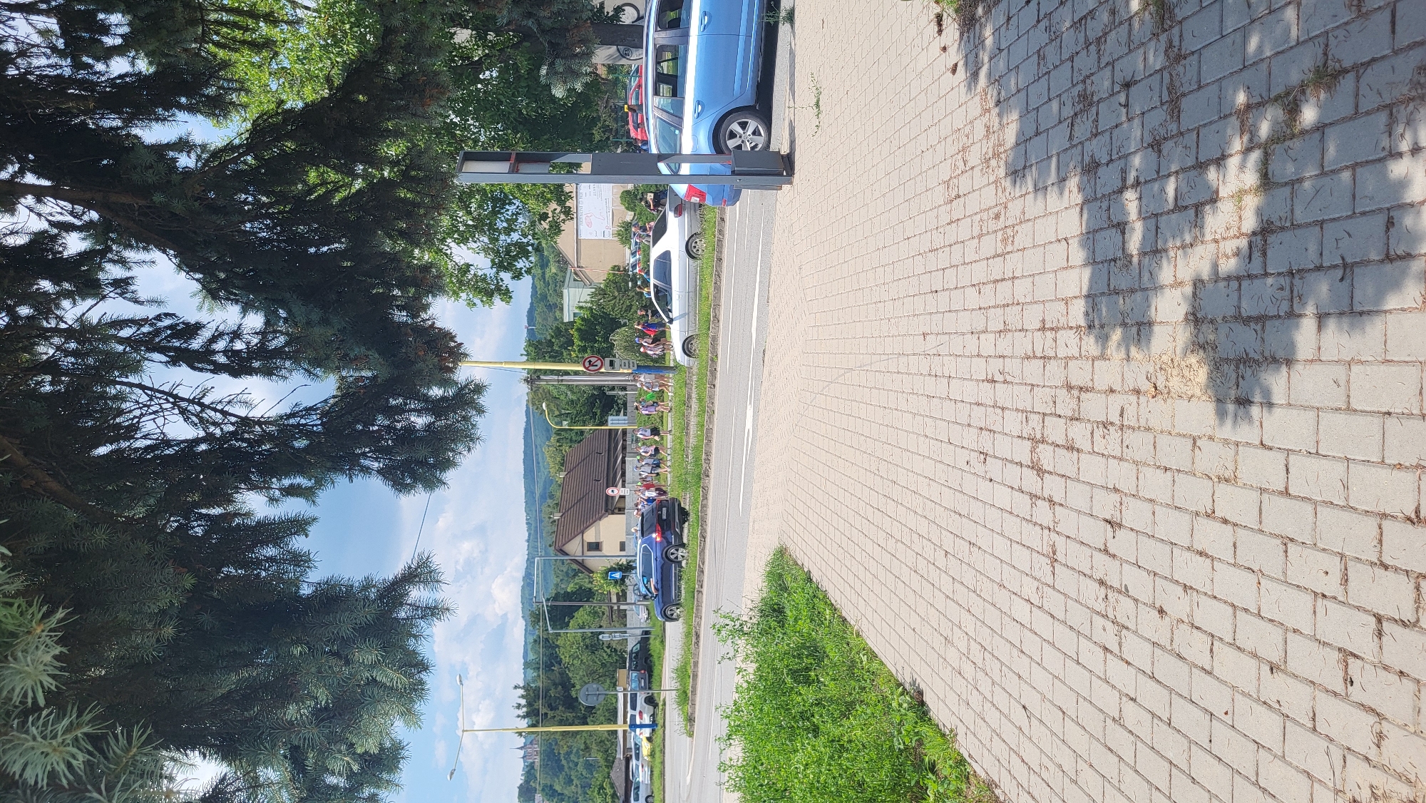 The image depicts a street scene in Prešov, Slovakia. A paved pedestrian walkway occupies the foreground, with scattered debris and leaf litter. Beyond the walkway, a road is visible with several vehicles. A blue SUV is driving on the road, and a silver sedan is parked on the side. A group of people, appearing to be walking together, are visible on the opposite side of the road. In the background, buildings, trees, and hills are present under a partly cloudy sky. Streetlights and traffic signs are also visible along the road. The overall impression is that of a residential or commercial area on a bright, sunny day.