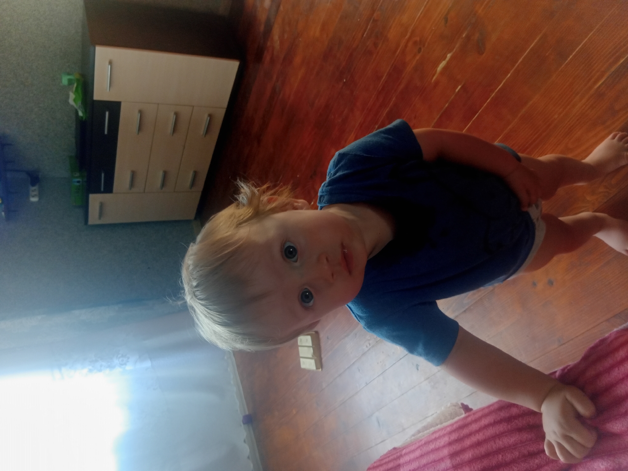 A young child with blonde hair and blue eyes, wearing a blue t-shirt and a diaper, stands on a wooden floor in what appears to be a room in Verkhniachka, Ukraine. The child is looking directly at the camera with a curious expression. To the left, a pink blanket is visible, and behind the child, a dresser with drawers and cabinets is against a patterned wallpapered wall. Sunlight streams in from a window on the left side of the image.