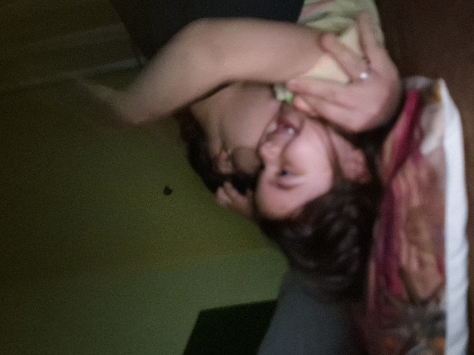 A person with dark hair is lying on a pillow with a pink and white patterned pillowcase. The person is looking towards the camera and sticking out their tongue. The person's arm is raised over their face, with fingers visible. The room appears to be indoors, with a green wall visible in the background. There is a dark object on the wall and a dark-colored object in the lower center of the frame. The exact location is difficult to discern from the image. The scene does not provide information related to Byala Slatina, Bulgaria.