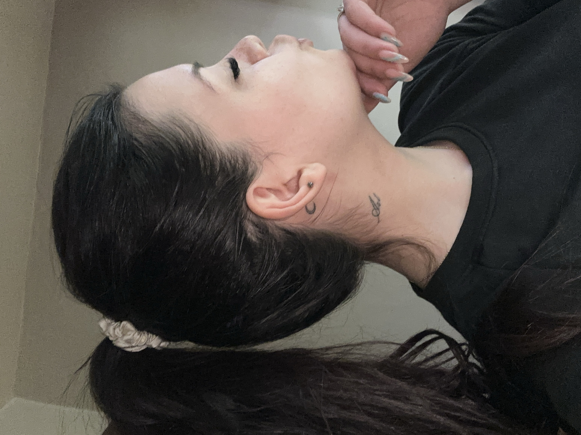 A close-up, profile shot depicts a woman with dark hair partially gathered in a ponytail with a light-colored scrunchie. She is wearing a black top. Her head is tilted back, exposing her neck, where a small tattoo is visible. A hand with long, manicured nails is near her face, seemingly touching her chin. She has a piercing in her earlobe. The background is out of focus, suggesting a neutral-toned wall. No evidence suggests the location is Sliven, Bulgaria.