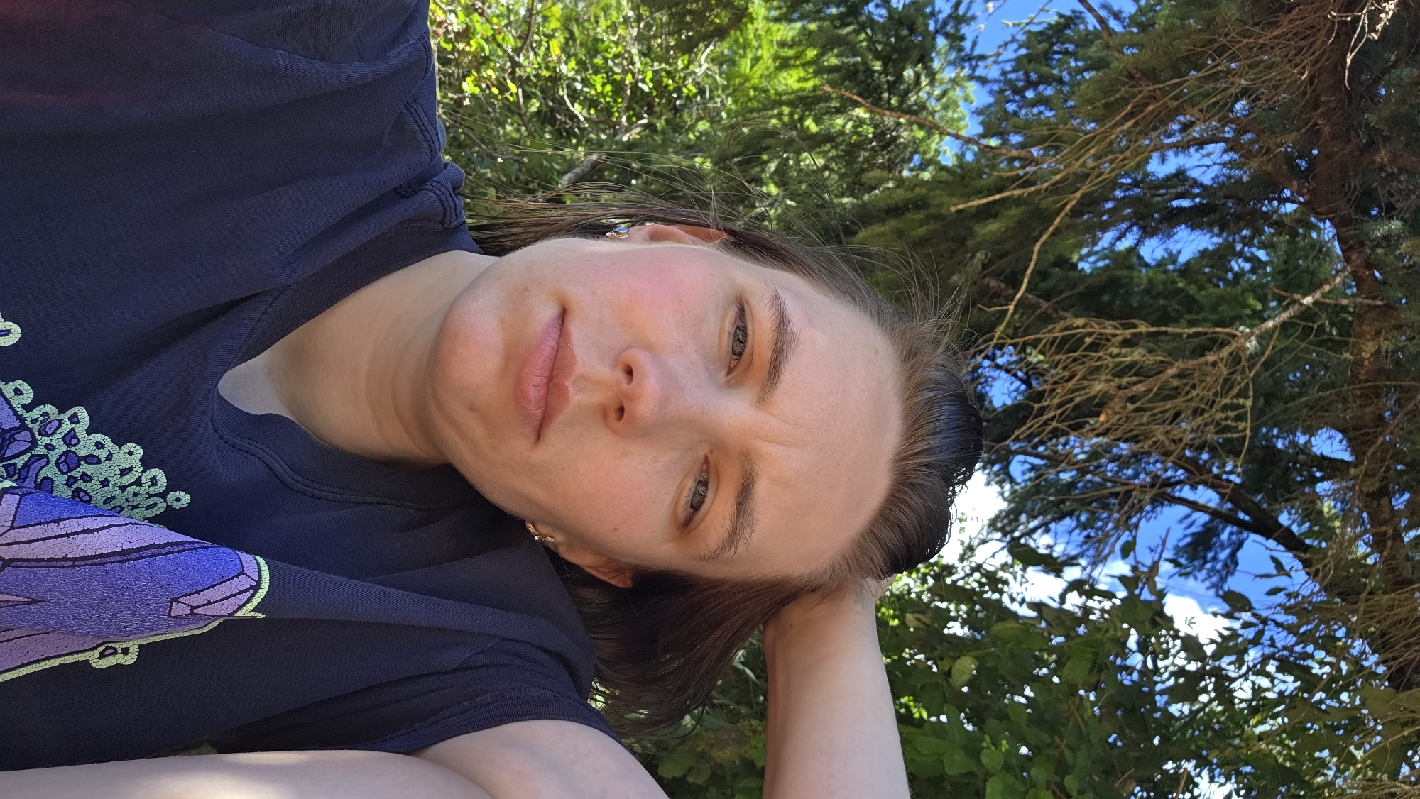 A woman is taking a selfie outdoors in what appears to be a park or wooded area. She has her hand behind her head and is looking at the camera with a slightly pensive expression. The background is filled with lush green foliage and branches of trees, with patches of blue sky visible through the leaves. She is wearing a dark blue t-shirt with a graphic design on the front. The lighting suggests it is daytime, likely a sunny day, with dappled light filtering through the trees. The exact location in Cherry Grove, United States, cannot be determined from the image.