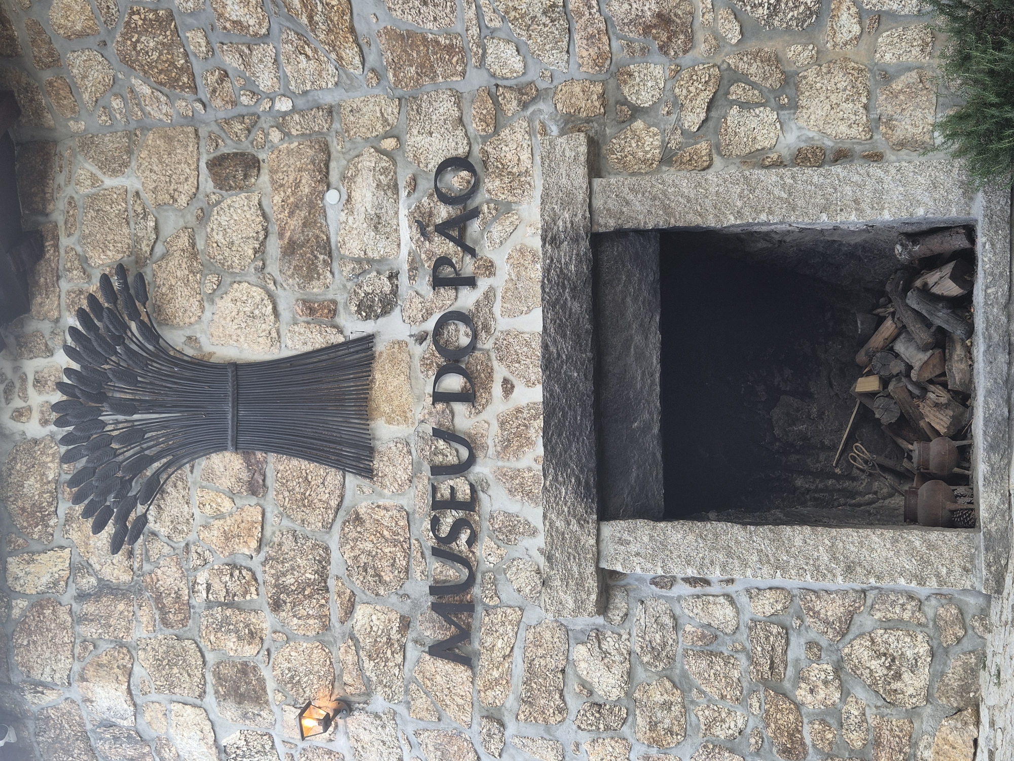 The image shows the exterior facade of the Museu do Pão (Bread Museum) in Seia, Portugal. The museum is built of rough-hewn, natural stone blocks of varying sizes, with visible mortar. Above a large, dark opening that appears to be a hearth or oven, black metal letters spell out "MUSEU DO PÃO". Directly above the lettering is a large, dark metal sculpture resembling a bundle of wheat stalks. To the left of the sculpture, mounted on the stone wall, is a single, old-fashioned outdoor lamp with an orange glow. The opening of the hearth contains firewood and what appear to be two cast-iron pots with legs. A sprig of rosemary or a similar herb is visible in the lower right corner of the image, against the stone wall. The lighting suggests it is daytime, possibly late afternoon or early evening given the warm glow of the lamp.