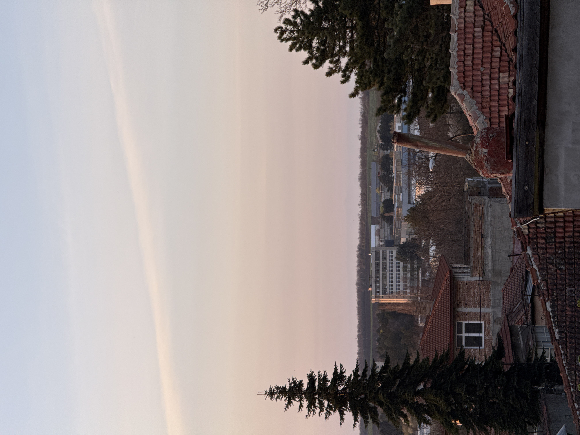 This media file presents a panoramic view of Dobrich, Bulgaria, captured during what appears to be late afternoon or early evening. The image, rotated 90 degrees clockwise from its intended landscape orientation, shows a sky transitioning from a pale blue on the left to a soft pinkish-orange on the right, indicative of a sunset or sunrise. A faint, elongated cloud or contrail stretches diagonally across the upper sky.
In the foreground and mid-ground, dense, dark green coniferous trees frame the scene from the top right and bottom. Amidst these trees and closer to the viewer are several residential buildings with characteristic red tiled roofs, some showing signs of age or partial dilapidation. One prominent detail is a brick chimney or similar pipe-like structure rising from among these older buildings. Another building, with a visible white-framed window against a brick wall, stands out.
Further in the distance, a larger, multi-story building with a modern appearance (possibly an apartment block or commercial structure) is visible, contrasting with the older architecture. Beyond the built-up area, the landscape flattens into what looks like fields or undeveloped land, stretching towards a hazy horizon. No specific activities or people are visible, lending a quiet, observational quality to the scene. There is no visible text in the image.