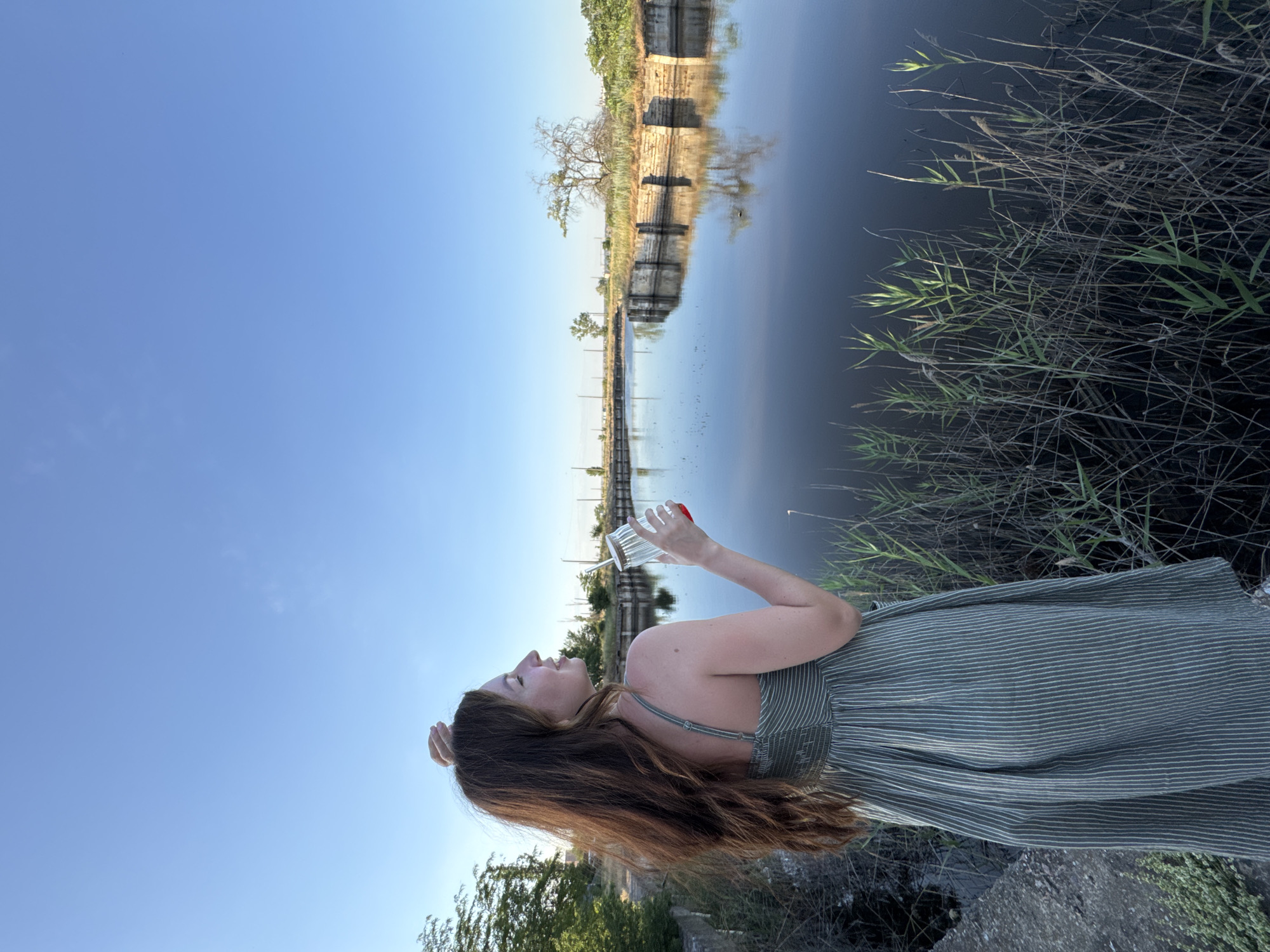 This vertically oriented, eye-level media file captures a young woman outdoors by a body of water. The woman, with long, brown hair, is wearing a green, pinstriped dress. She is holding a clear glass with a straw and a red lid, appearing to drink from it. Her head is tilted back, and her eyes are closed, suggesting a moment of enjoyment or relaxation.
The scene is a waterfront, with dark, calm water filling a significant portion of the frame. The water reflects the sky and a man-made structure that lines the opposite bank. This structure appears to be a concrete embankment with stone accents and is bordered by vegetation and trees. Tall, dry grasses and reeds grow along the water's edge in the foreground and right side of the image. The sky is a clear, bright blue, indicating a sunny day. The lighting suggests it is either early morning or late afternoon.
The overall mood is peaceful and serene. The location cues, such as the vegetation and the type of waterway, are consistent with a natural or semi-natural environment. There is no visible text in the media file.